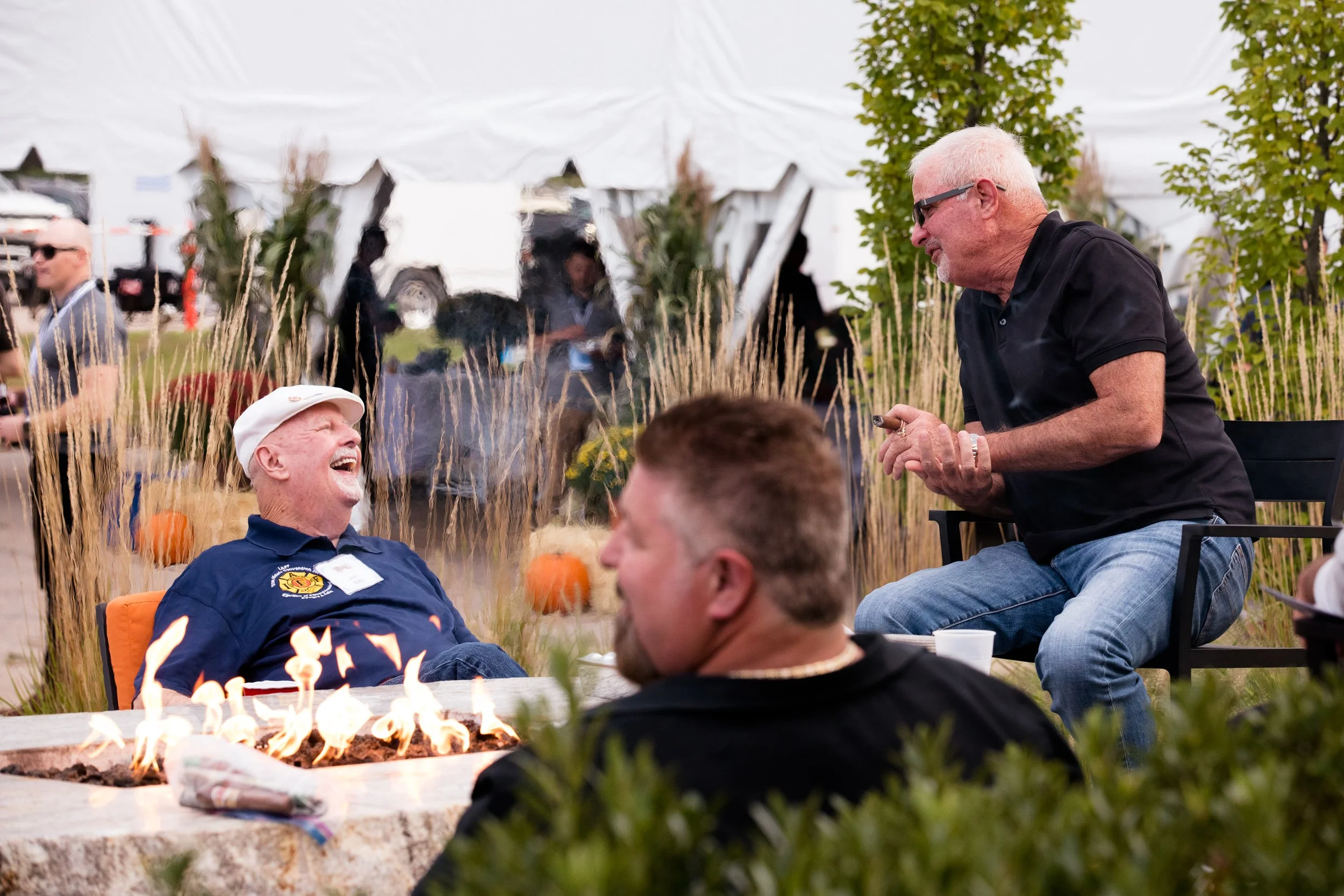 People enjoying a gathering outdoors around a fire pit, with one man laughing and another man holding a cigar.
