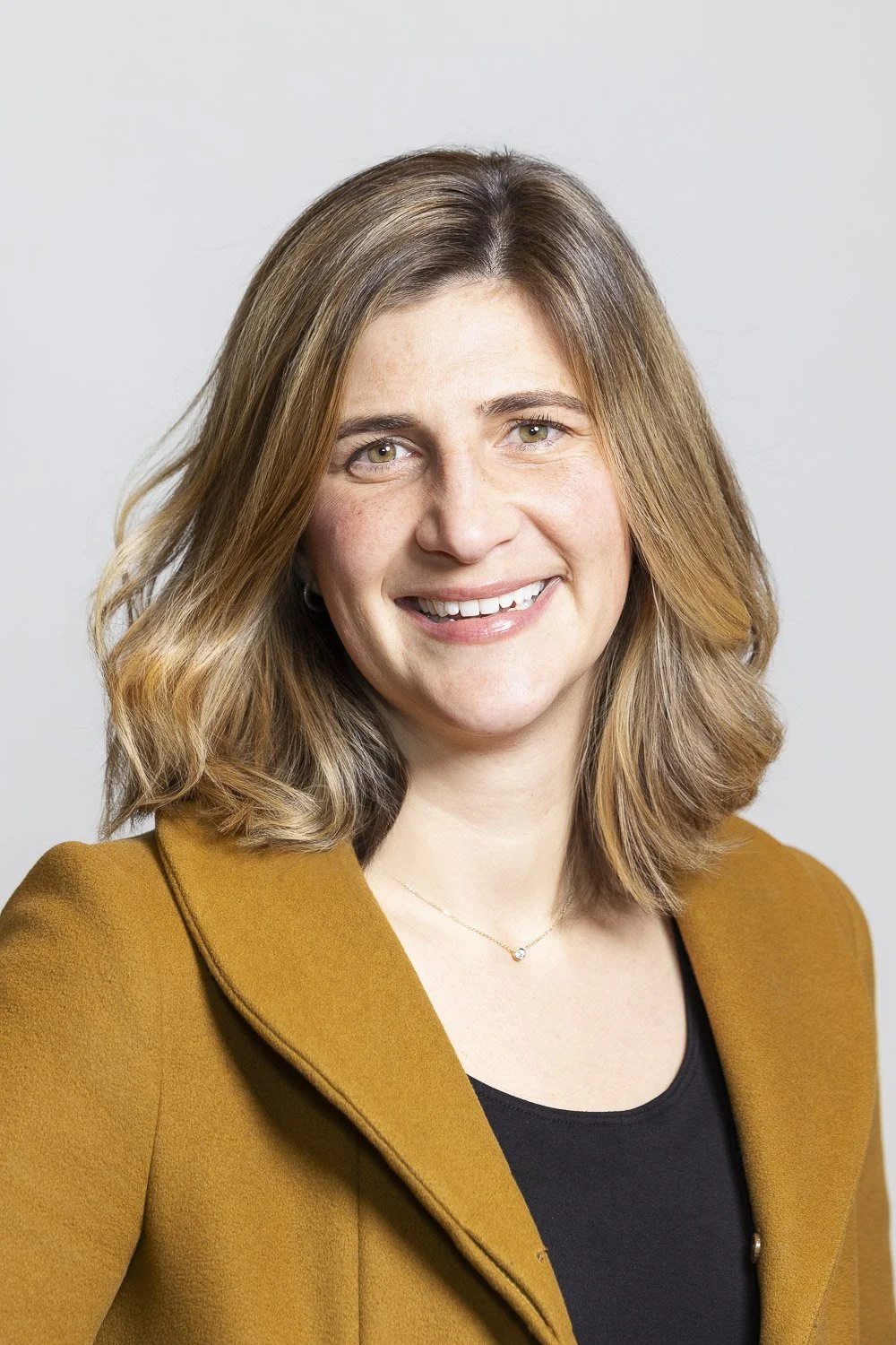 A smiling woman wearing a mustard-colored blazer and a black top, against a plain light background at am alumni networking event in Boston, Massachusetts.