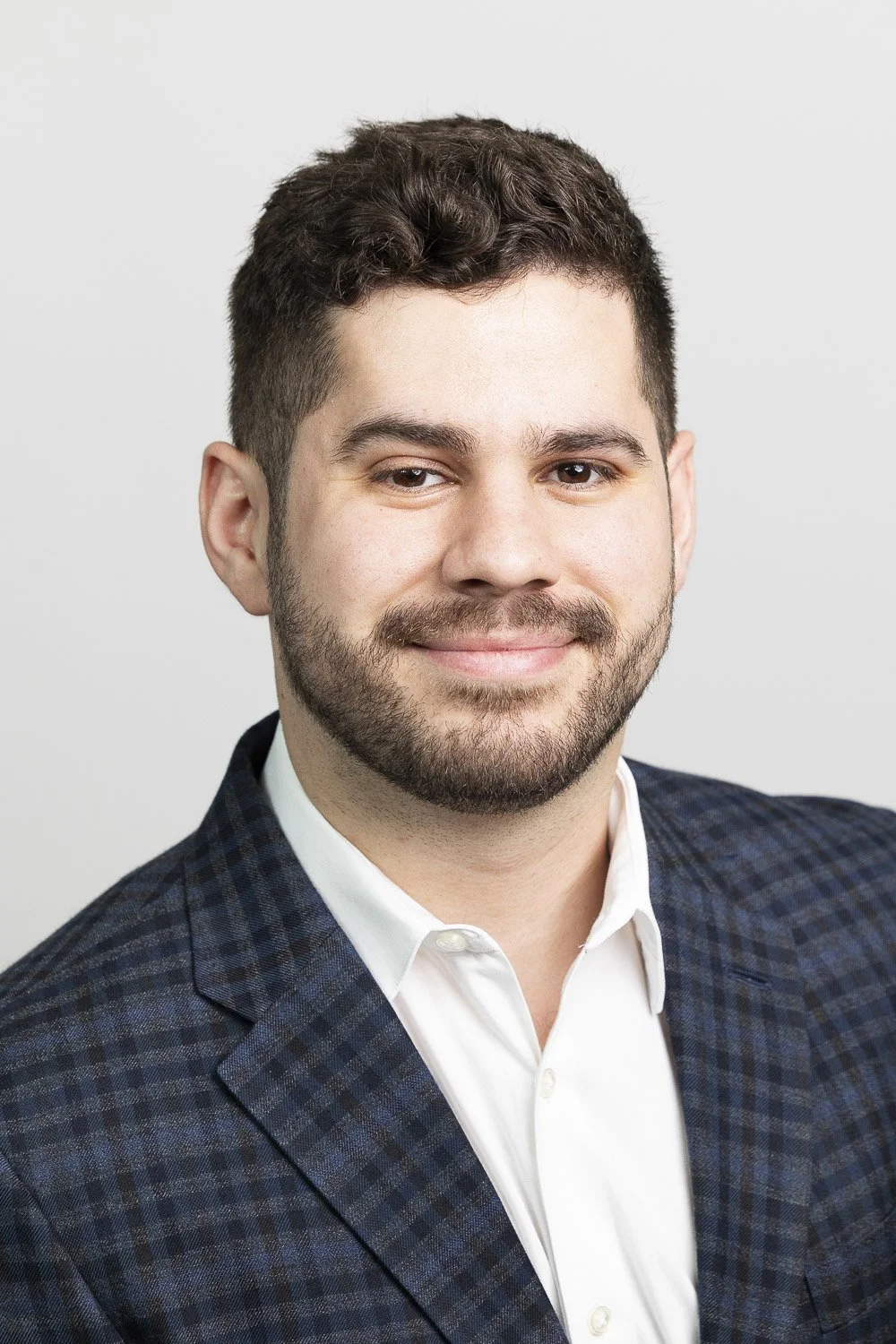 A young man with short dark hair, a beard, and wearing a blue checkered blazer, smiling against a plain light gray background.
