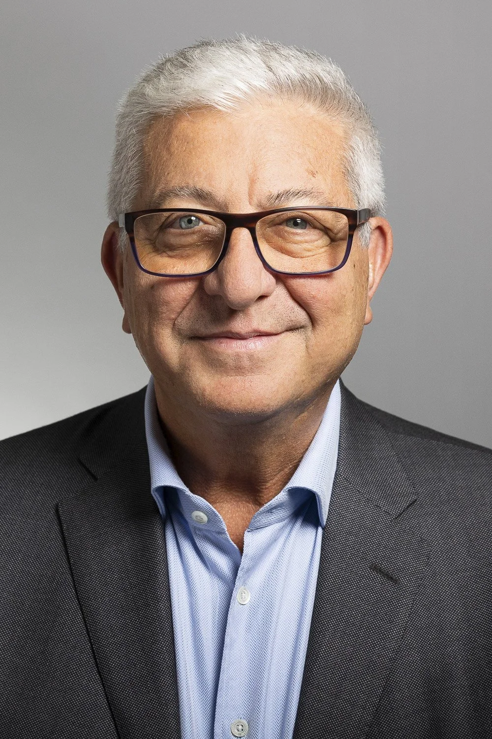 Headshot of an older man with gray hair and glasses, wearing a dark blazer and light blue dress shirt, smiling against a plain gray background.
