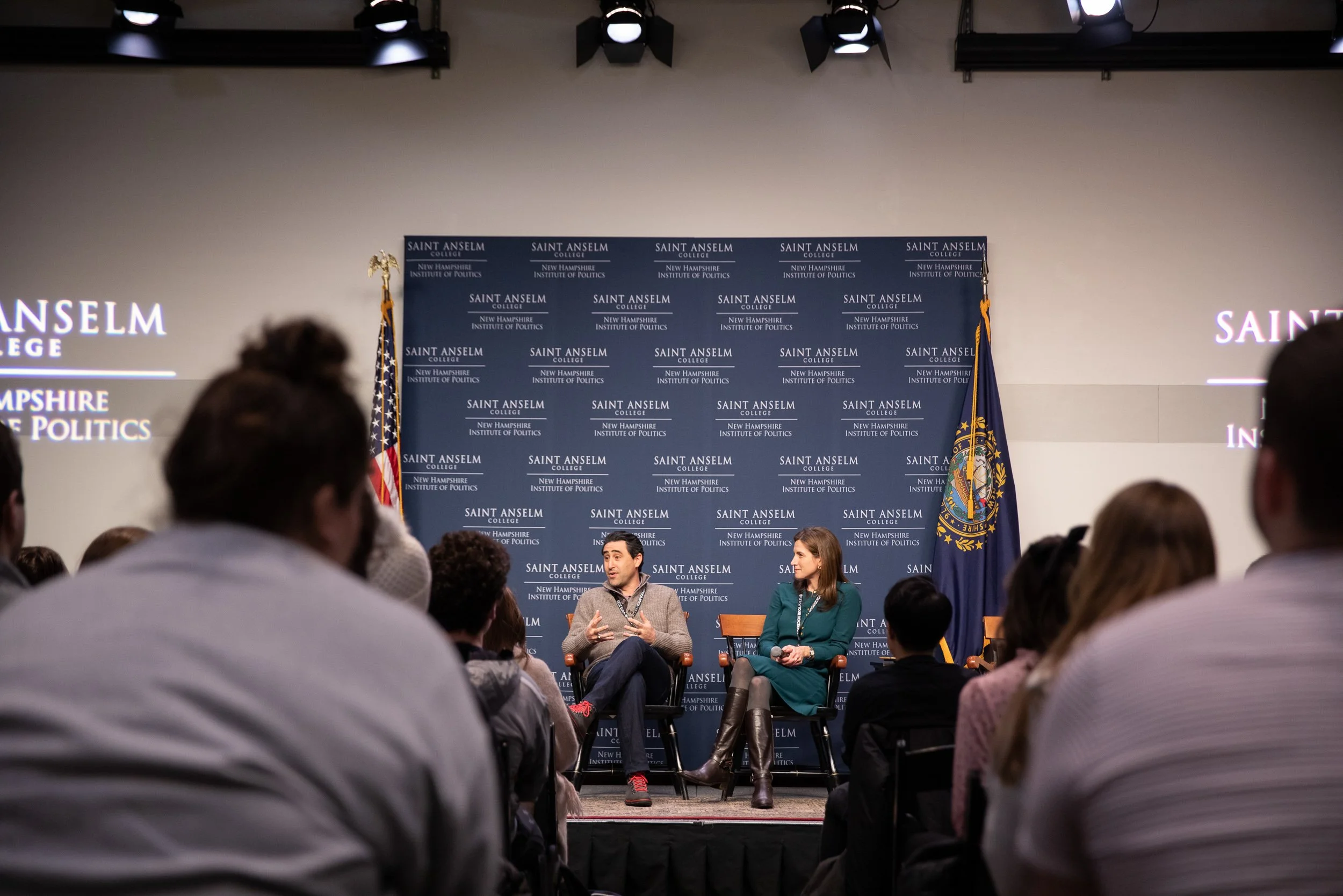 A speaker and a moderator seated on stage in front of an audience during an event at Saint Anselm College.