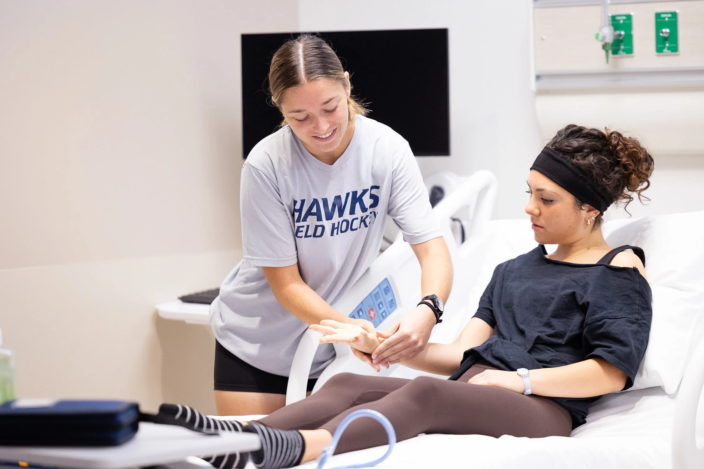 A nursing student helping another student in a clinical simulation setting.