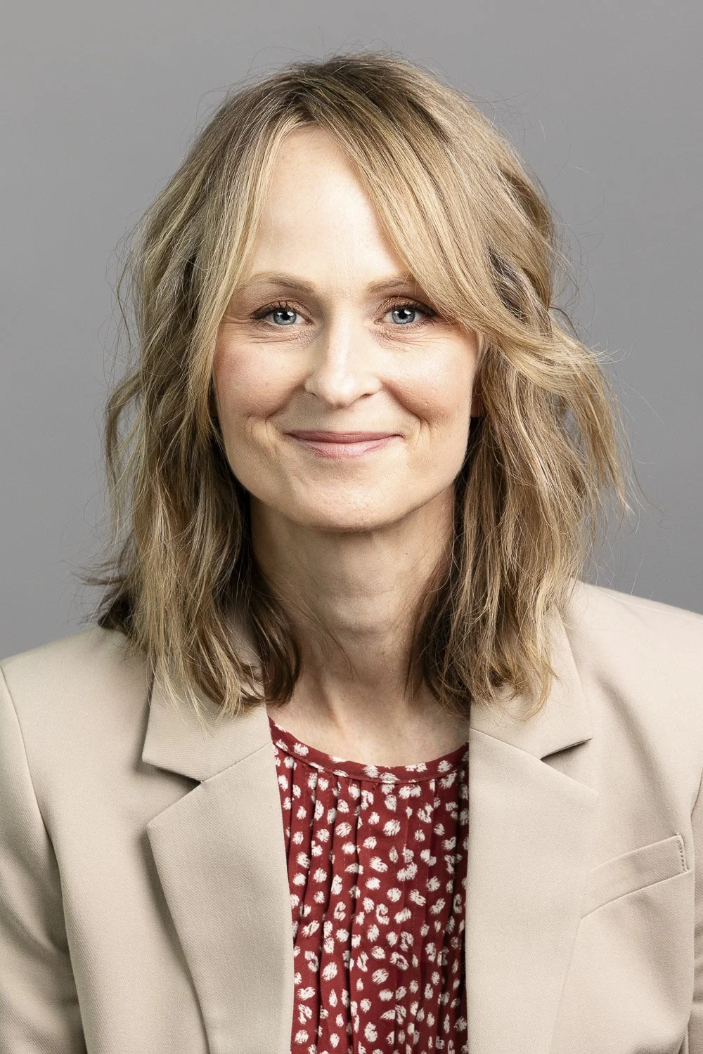 Headshot of a woman wearing a beige blazer over a red patterned blouse, smiling in front of a gray background at a studio in Massachusetts.