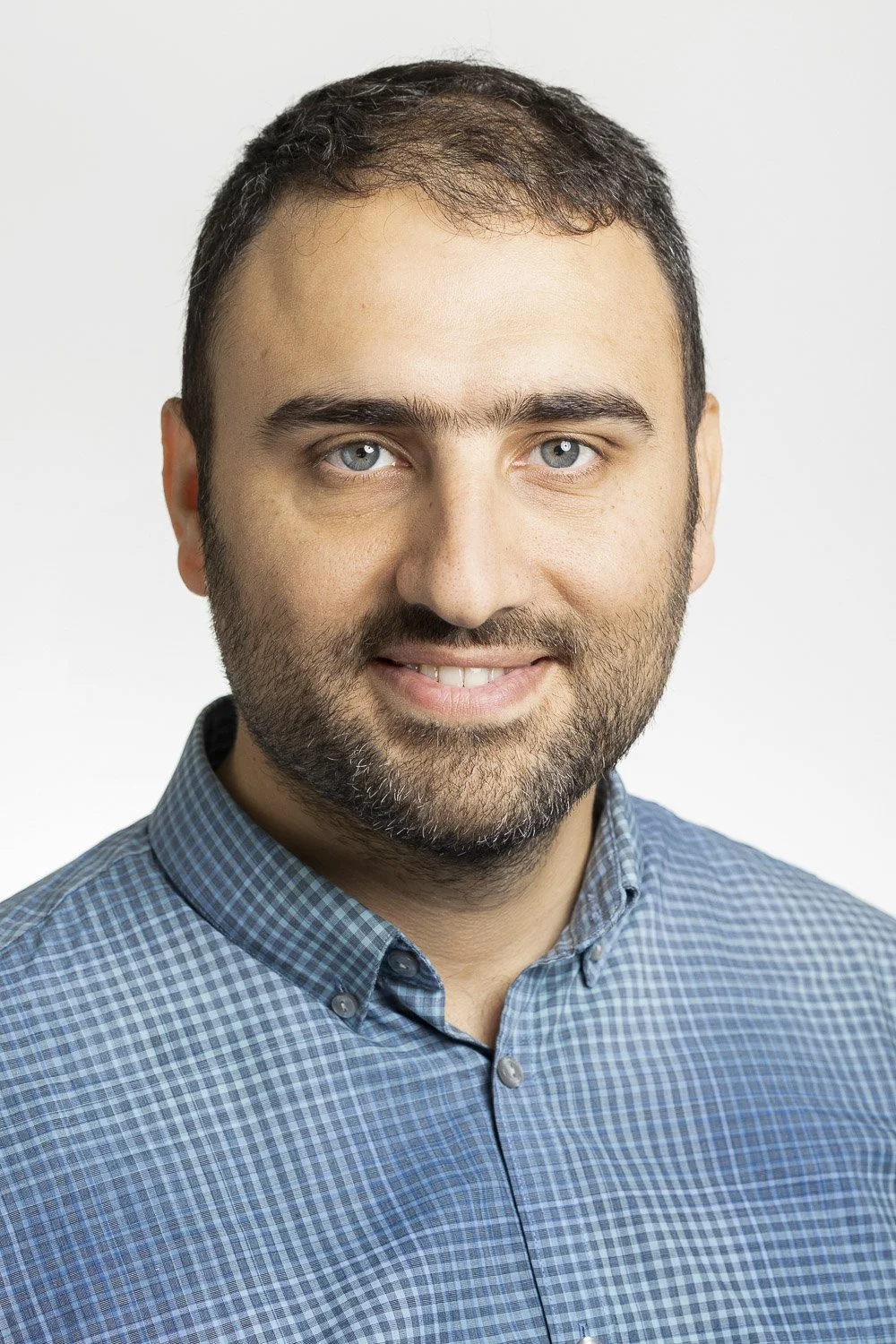 Portrait of a man with short dark hair, wearing a blue checked shirt, smiling, against a plain white background.