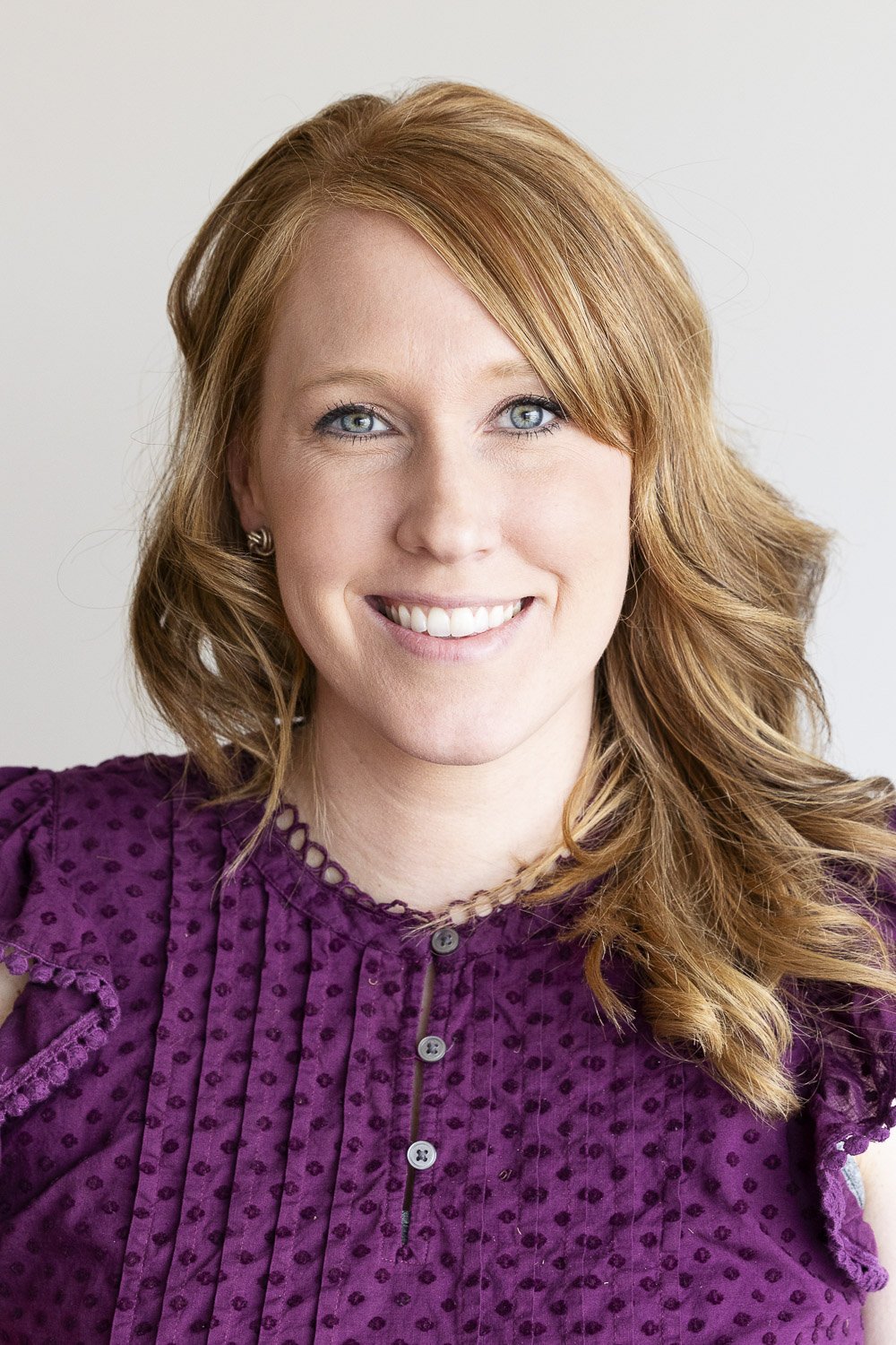 Portrait of a smiling woman with shoulder-length red hair, blue eyes, wearing a purple blouse with a textured pattern and ruffled details.
