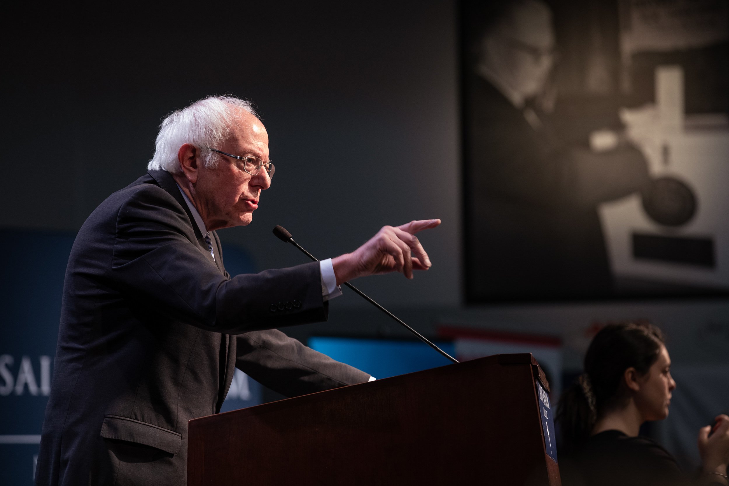 Bernie Sanders speaking at a podium, gesturing with his right hand, during a speaking engagement at Saint Anselm College.
