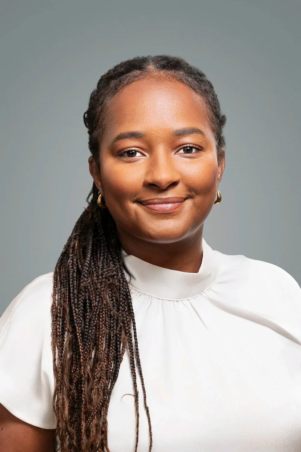 A professional woman wearing a white top and gold earrings, smiling against a grey background.