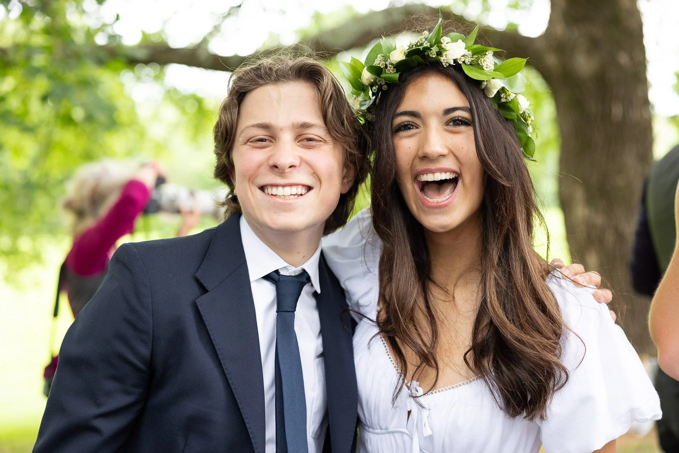 A young man in a suit and a young woman wearing a flower crown smiling and posing together outdoors. 