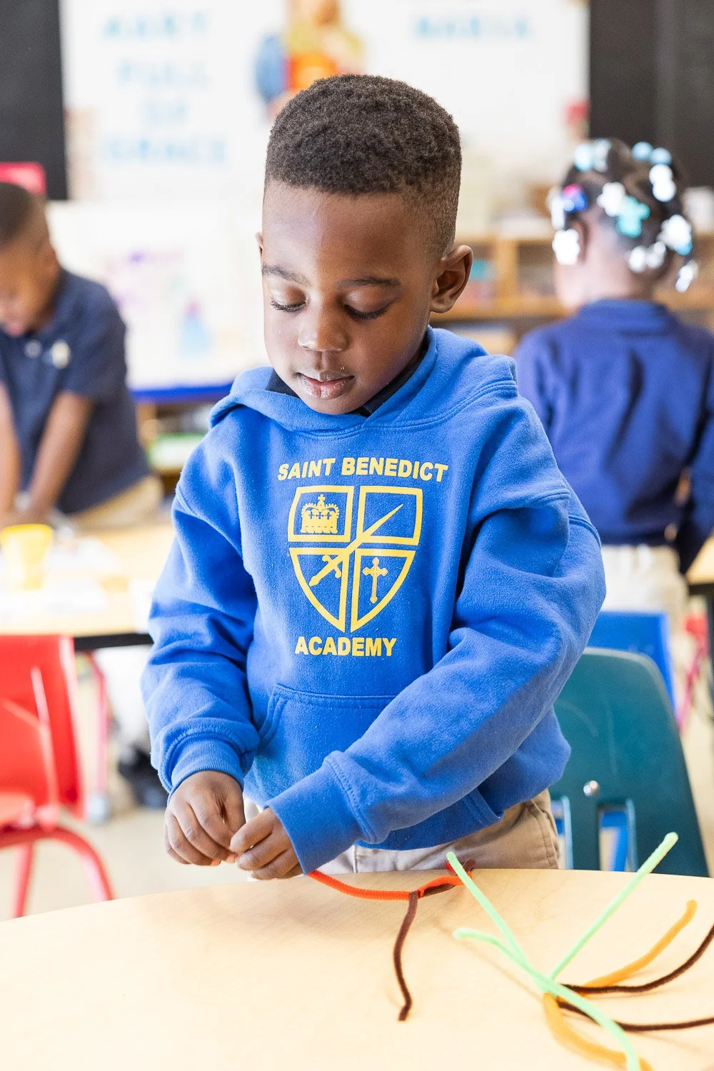 A young boy at Saint Benedict Academy engaged in a craft activity with pipe cleaners on a table.