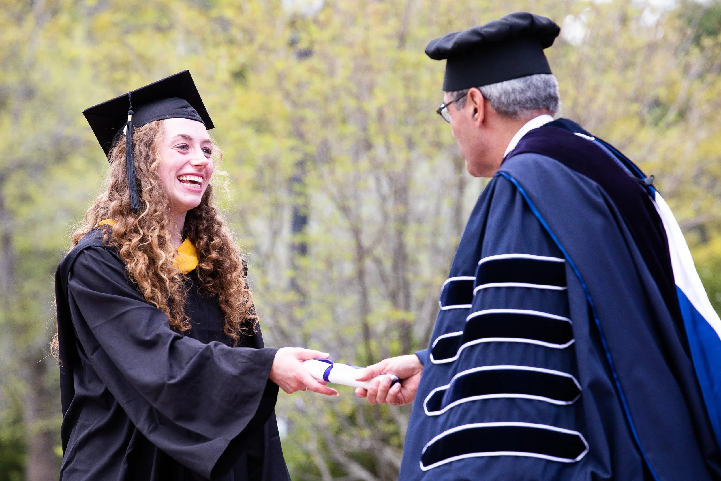 A young woman in a graduation gown and cap receiving her diploma from a man in academic regalia at an outdoor graduation ceremony.