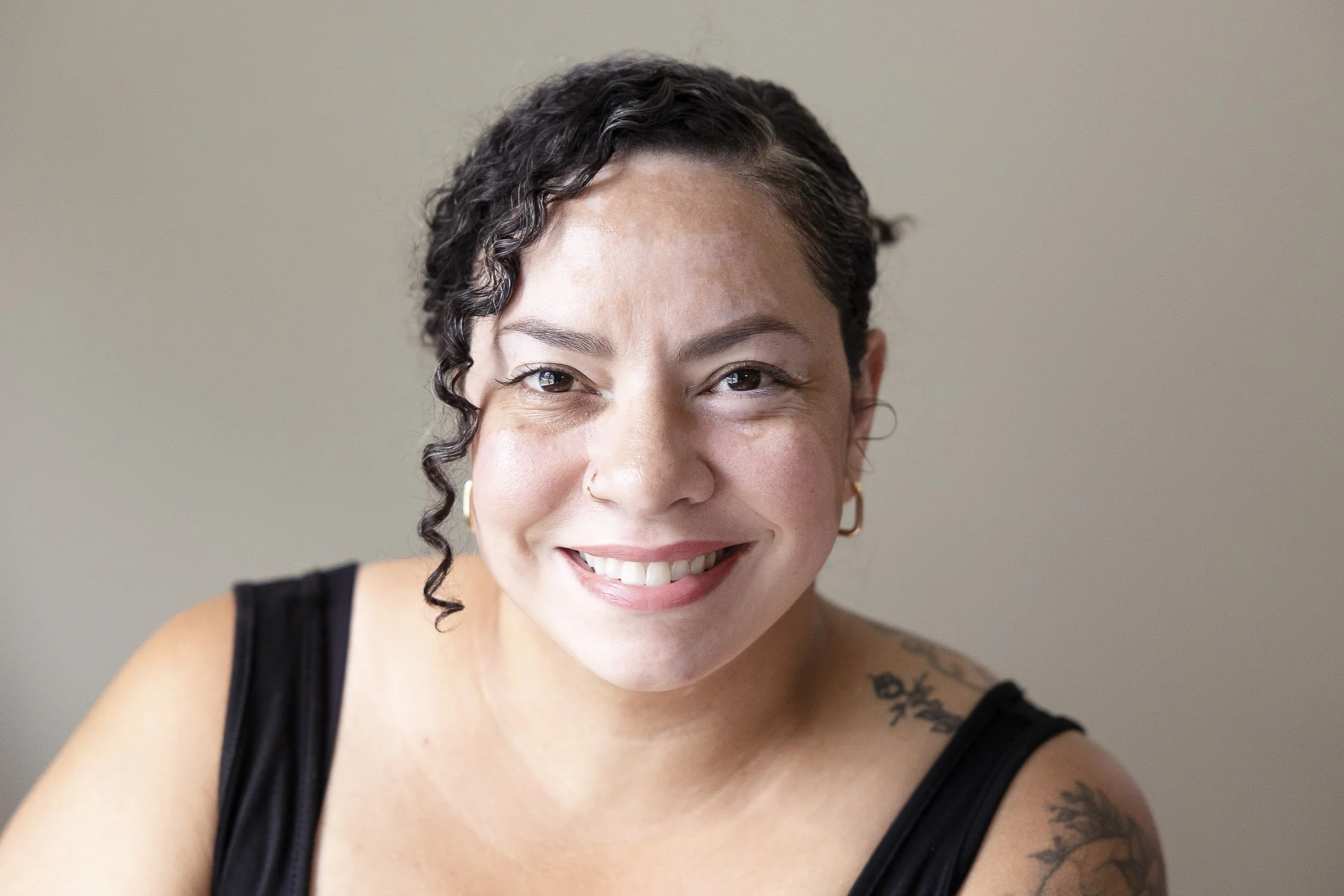 Close-up portrait of a smiling woman with short curly dark hair, wearing gold hoop earrings and a black tank top,.