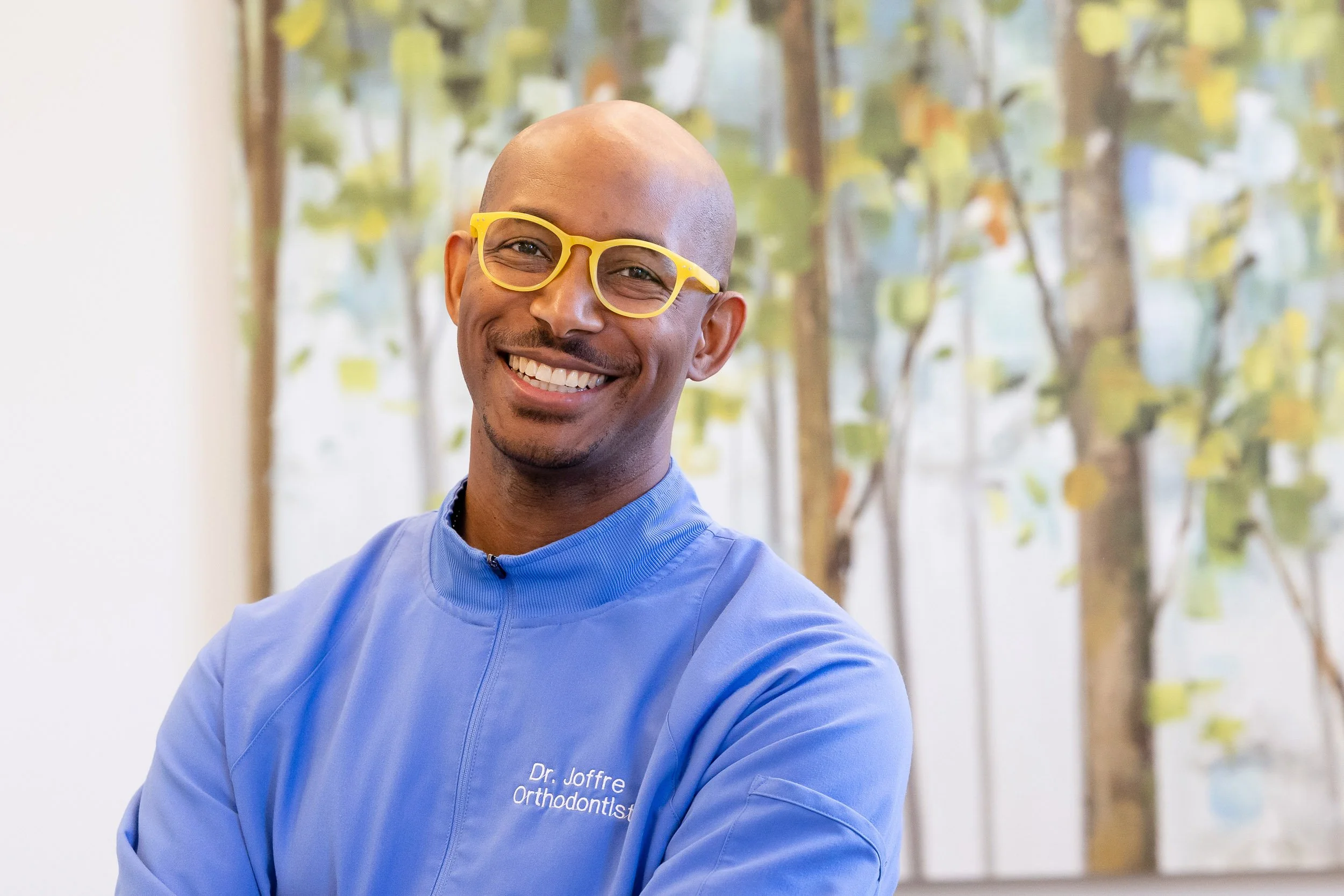 A smiling man wearing yellow glasses and a blue dental coat, standing in front of a colorful abstract background.