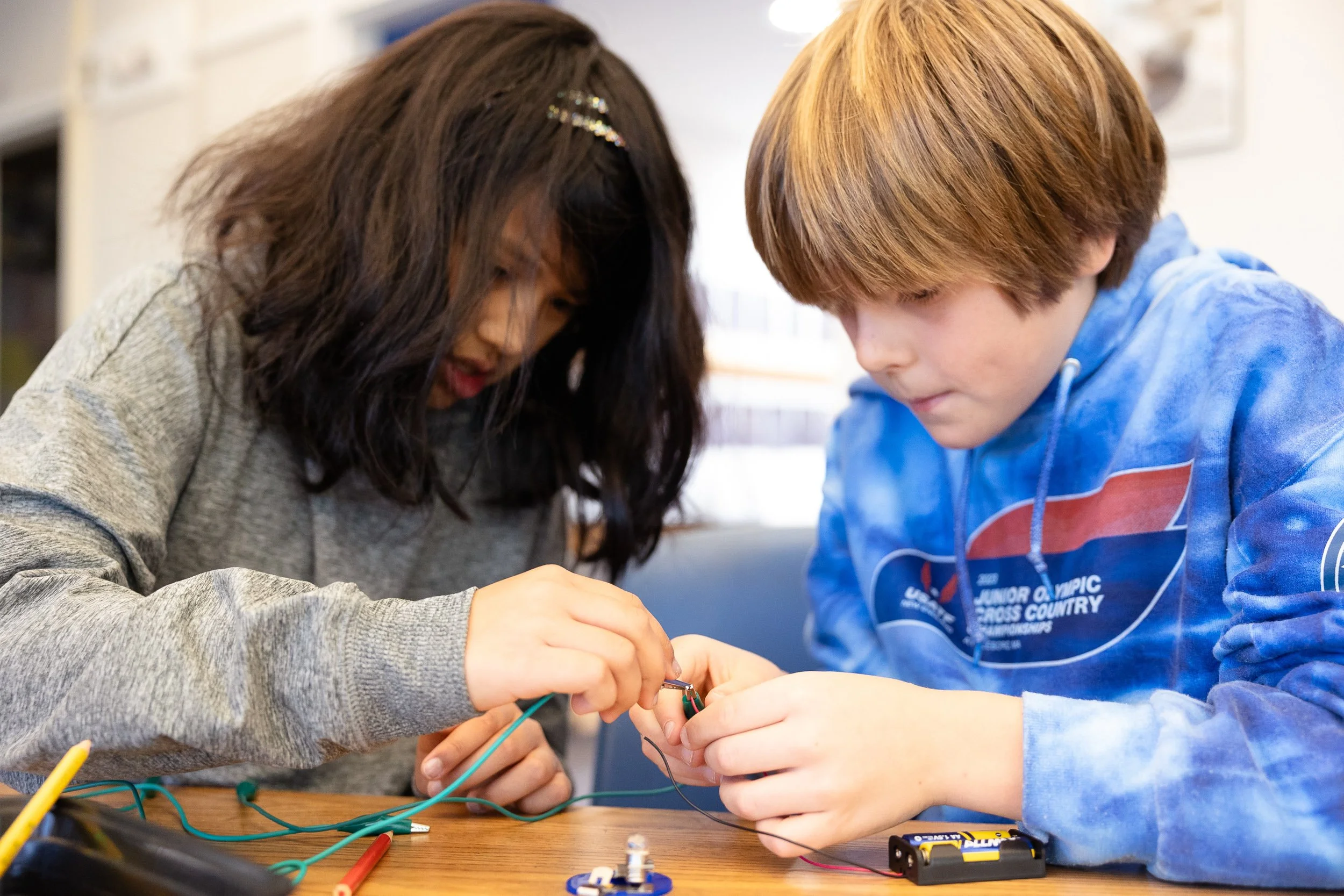 A girl and a boy are working together on an electronics project at a classroom table, connecting wires and components.