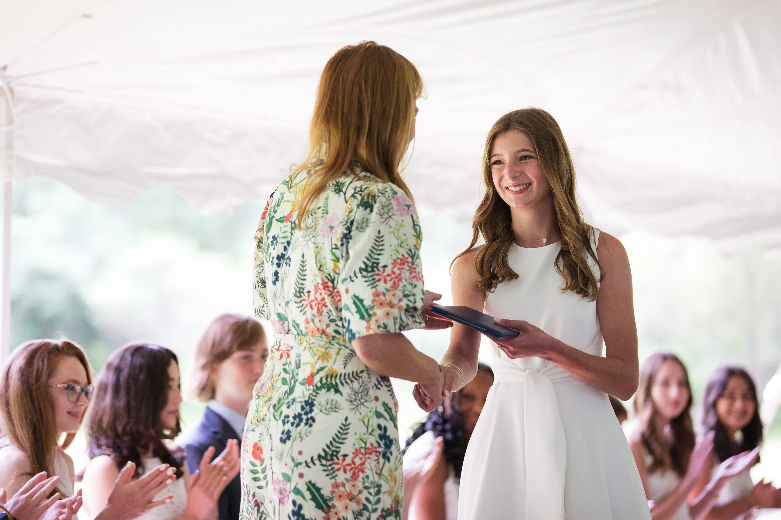 A young woman in a white dress receiving diploma in outdoor private school graduation ceremony.