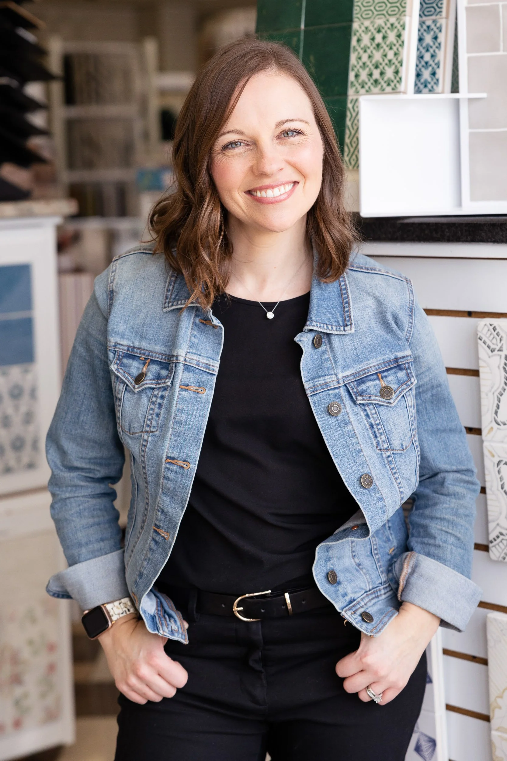 A smiling female interior designer with shoulder-length brown hair, wearing a denim jacket over a black top, black pants, standing inside a tile store in Natick, Massachusetts.