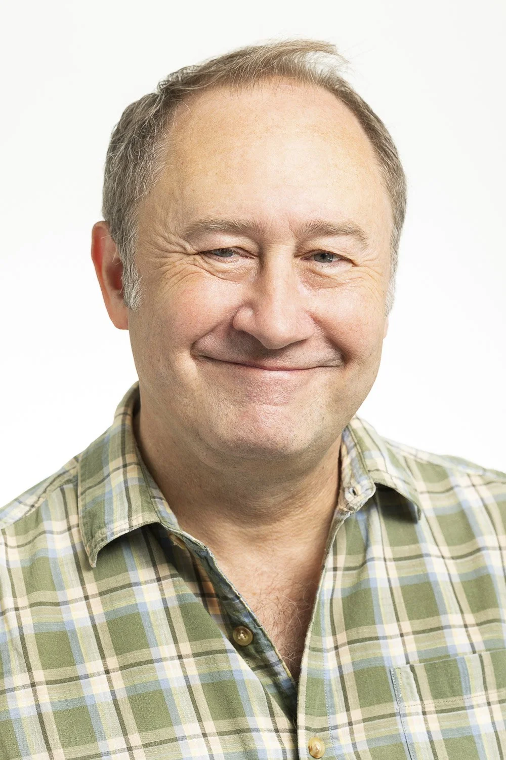 A middle-aged man with short, light-colored hair, smiling, wearing a plaid shirt, against a plain white background.
