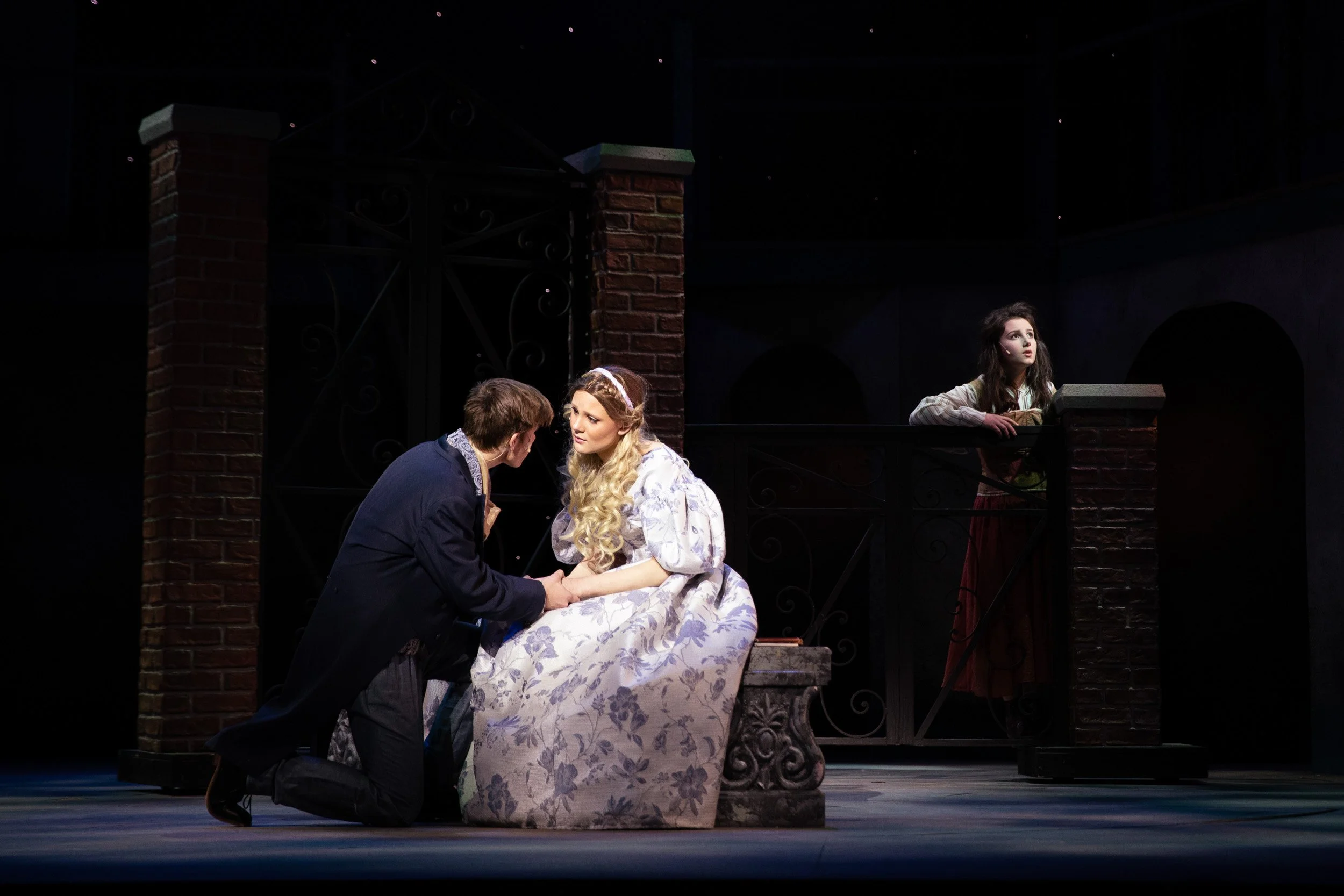 High school actors in a scene, one kneeling and holding hands with a woman in a long floral dress.