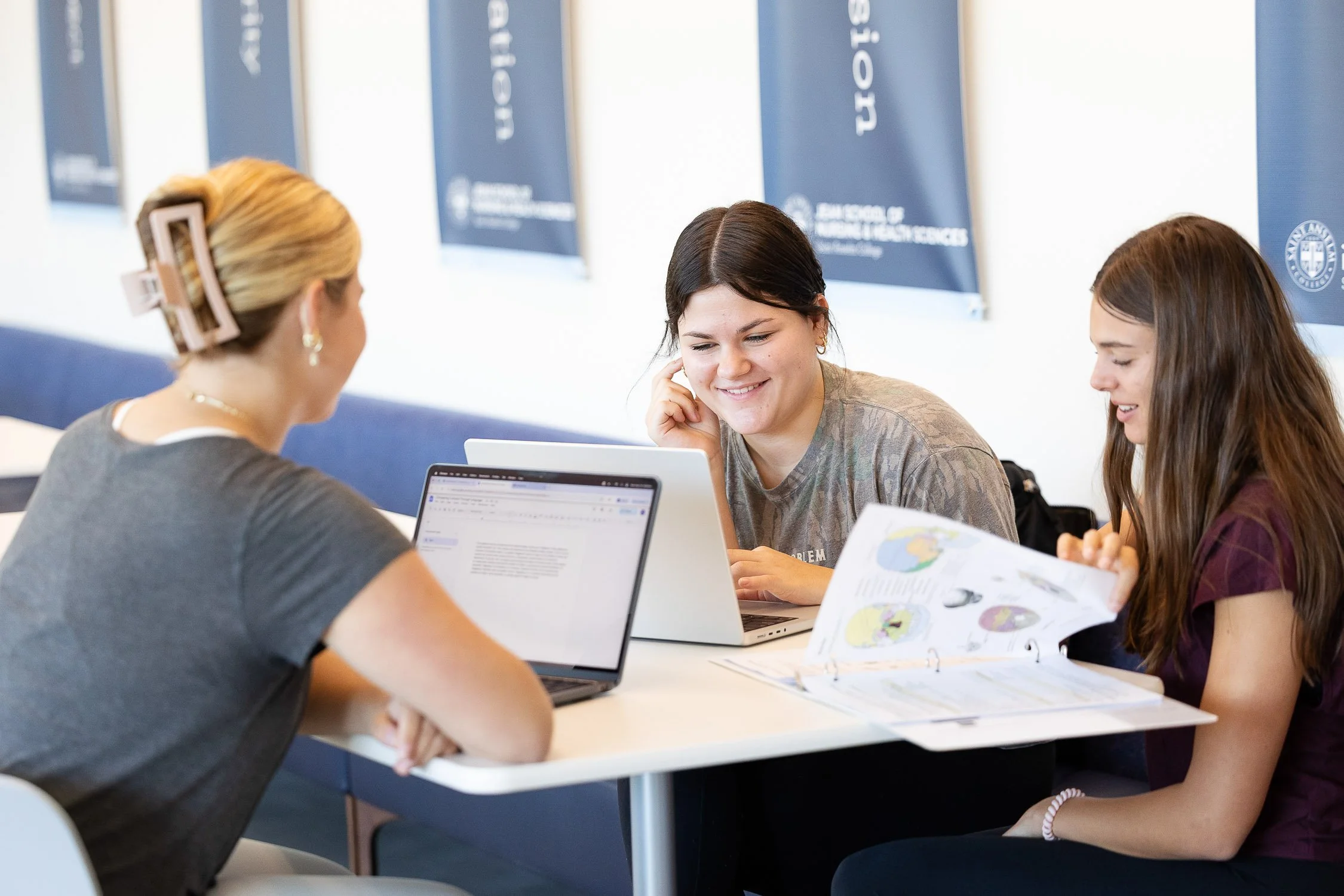 Three female students sitting at a table, working on laptops and looking at notes, on a college campus in New Hampshire.