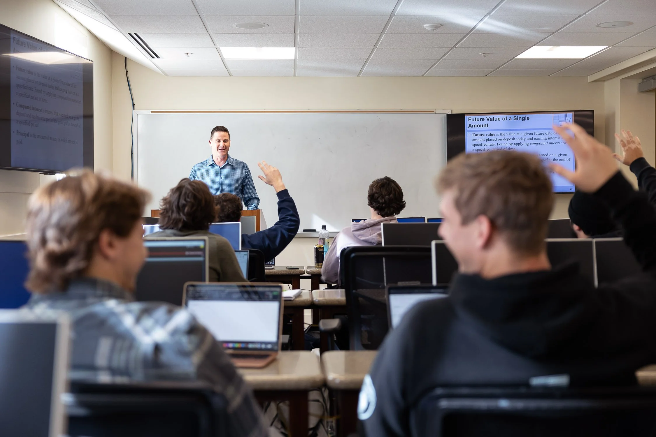 A college classroom with students at desks and laptops raising hands while a male teacher stands at the whiteboard.