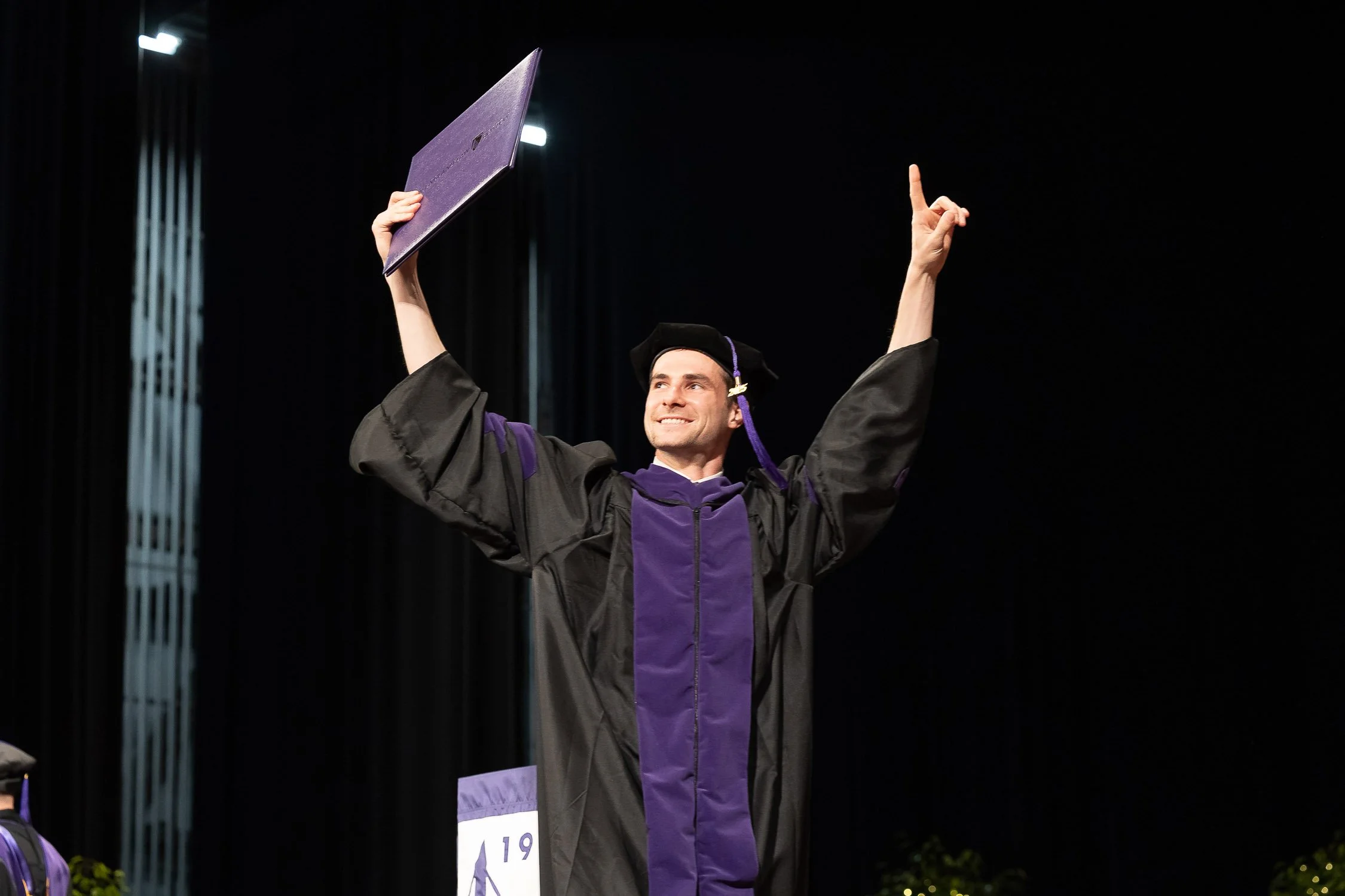 A man in graduation regalia holding a diploma in one hand and raising the other hand with an index finger up, smiling on stage during a graduation ceremony from law school in Boston, Massachusetts.
