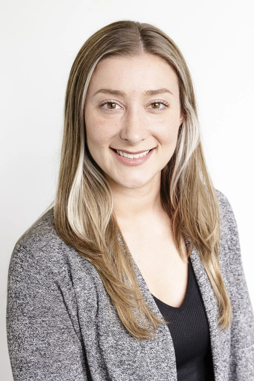 Portrait of a smiling young woman with long blonde hair, wearing a gray sweater and a black top, against a white background.