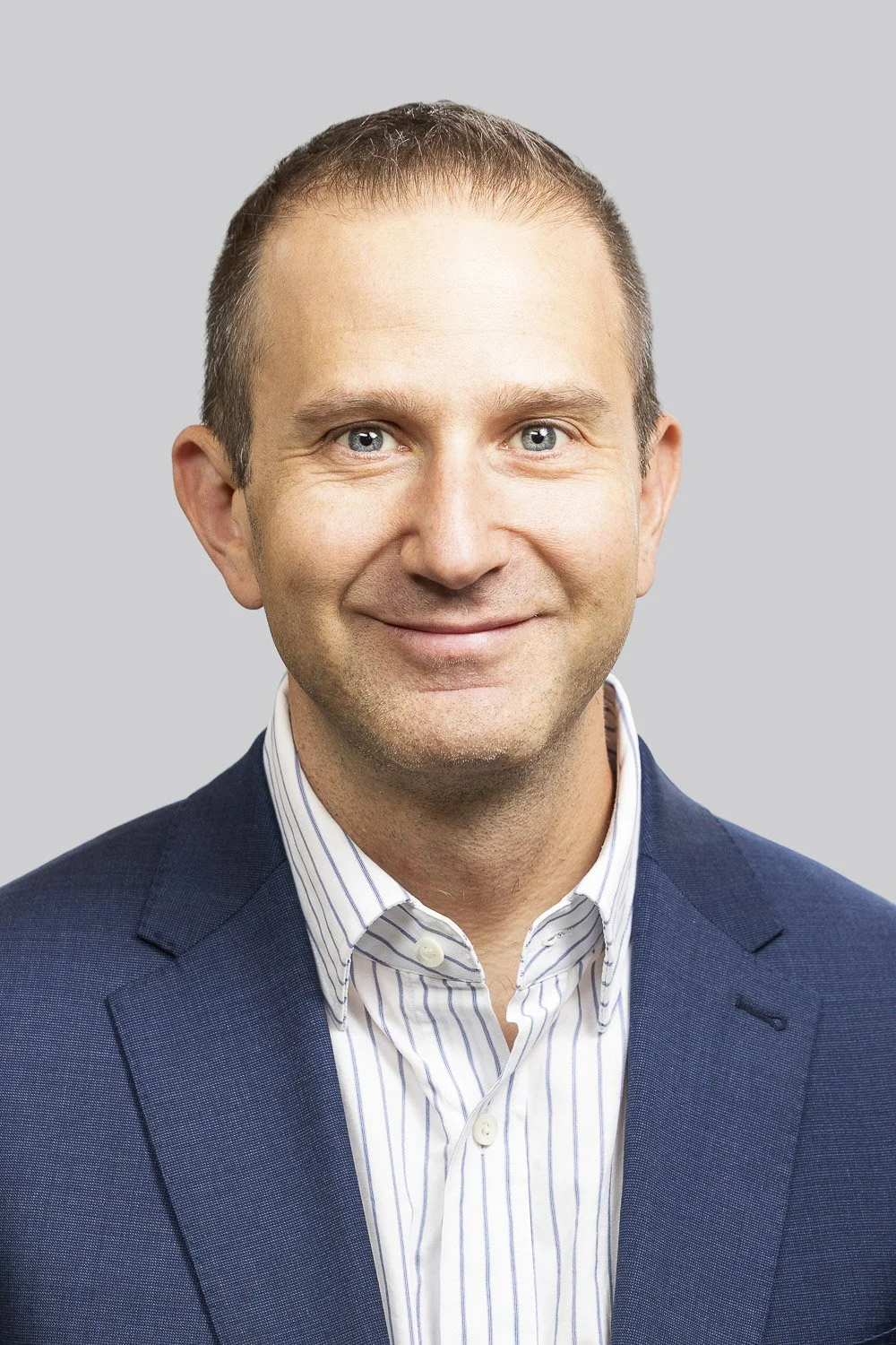 Headshot of a smiling man with short brown hair, blue eyes, wearing a navy blazer and a white striped dress shirt against a light gray background.