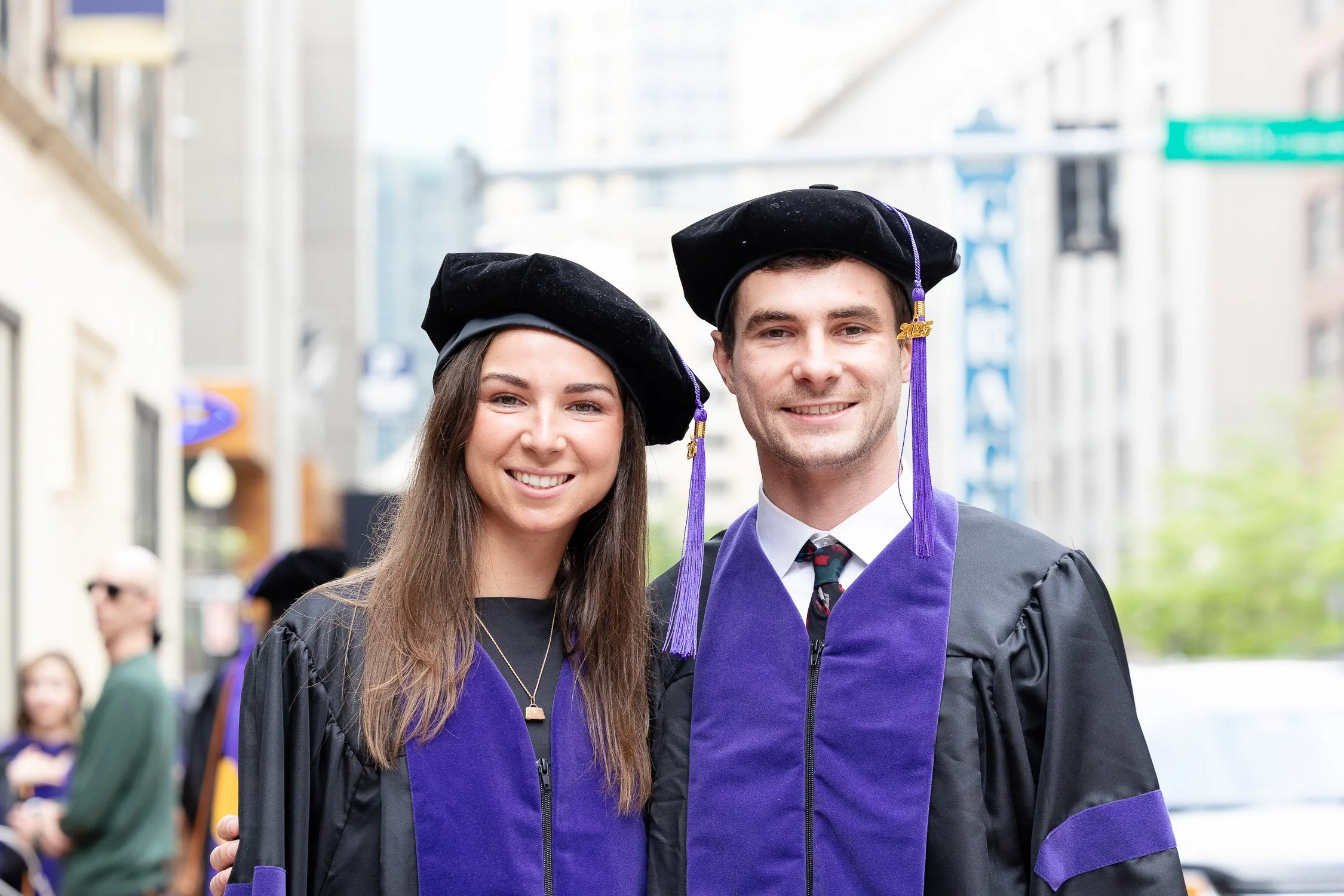 Two graduates in caps and gowns smiling at the camera, standing outdoors in Boston.