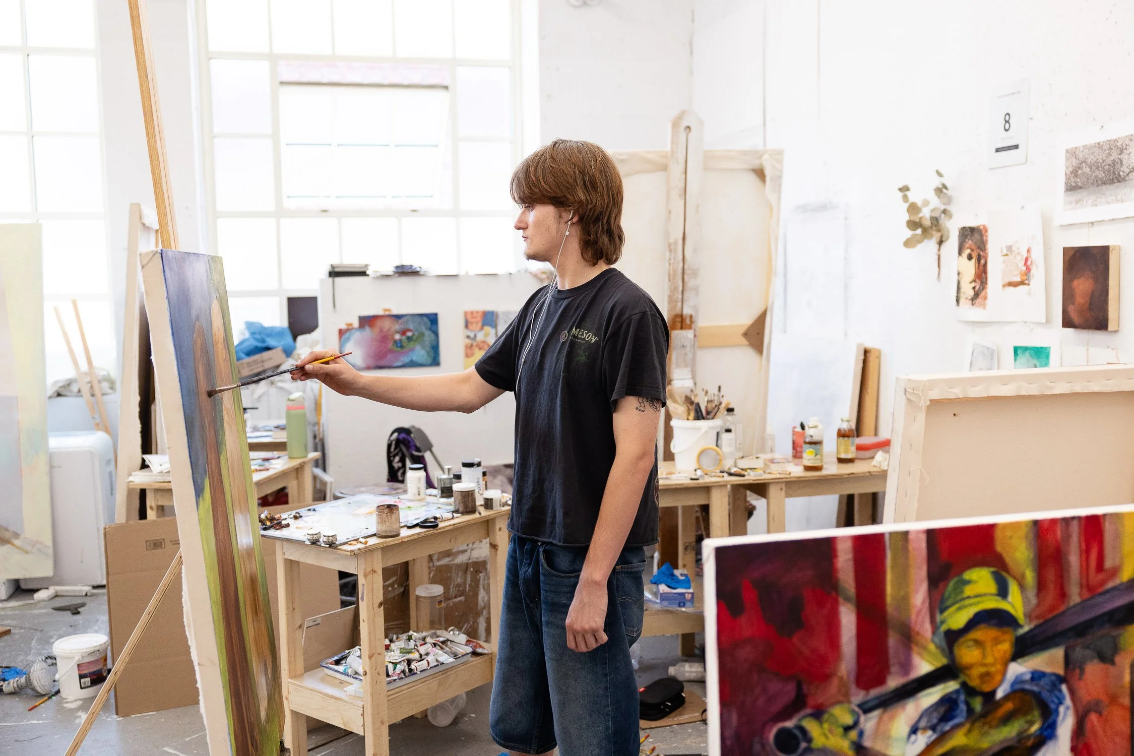 A student painting on a large canvas in an art studio with various paintings and art supplies around.