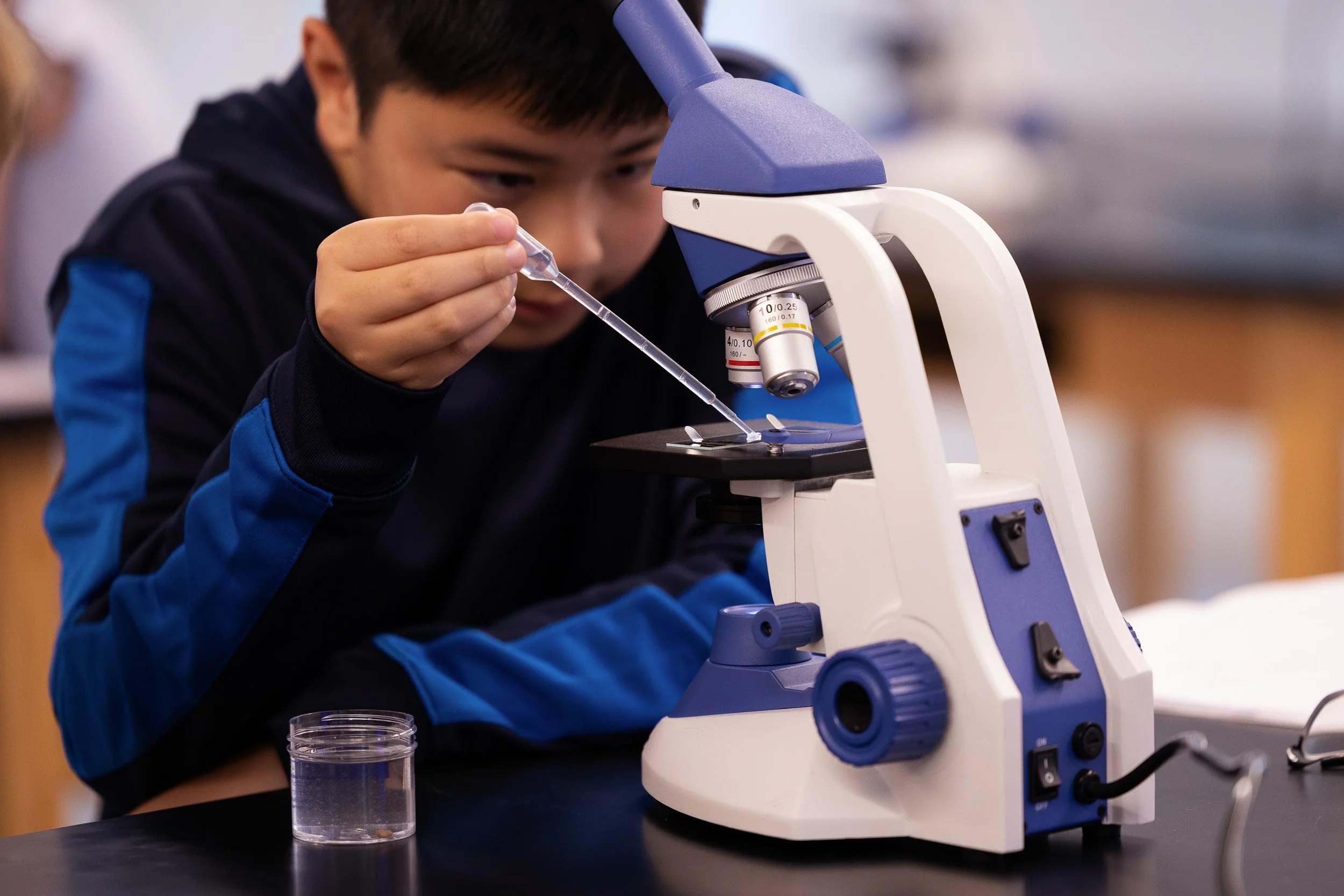 A middle school boy using a pipette to transfer liquid into a slide under a microscope in a science lab at a private school in Massachusetts.