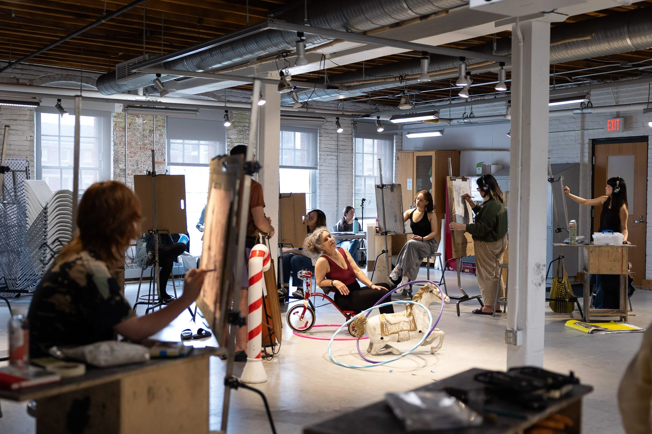 Artists working on a collaborative art project in a spacious art school studio with large windows, chairs, and various art supplies.