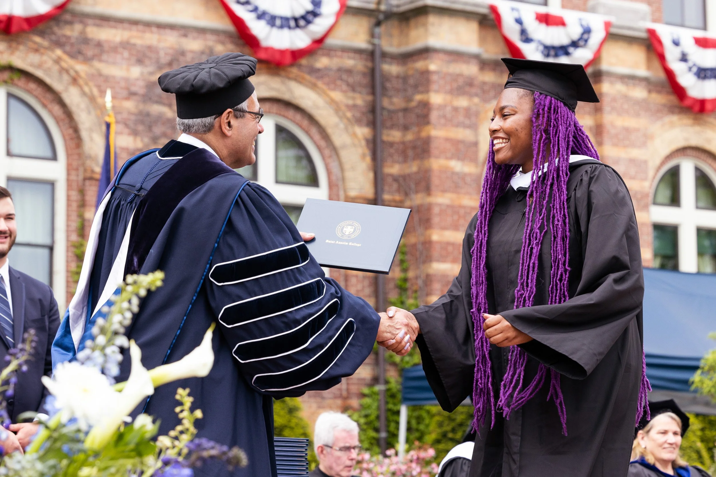 A woman in a black graduation gown and tassel receives her diploma from a man in academic regalia during a graduation ceremony outside a brick building at a college in Boston, Massachusetts.