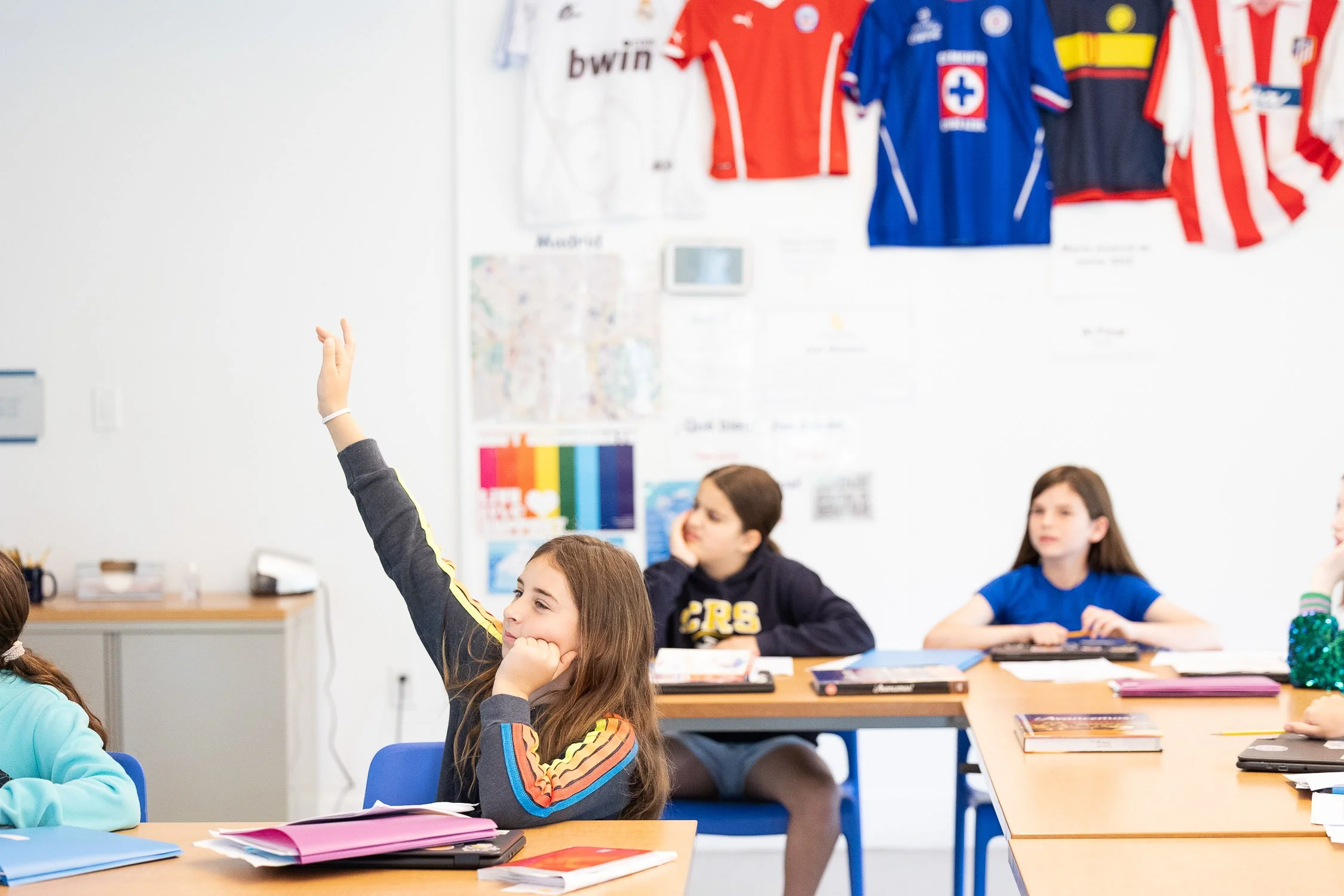 A girl in a classroom raising her hand while sitting at a desk with notebooks and papers at a Boston private middle school.