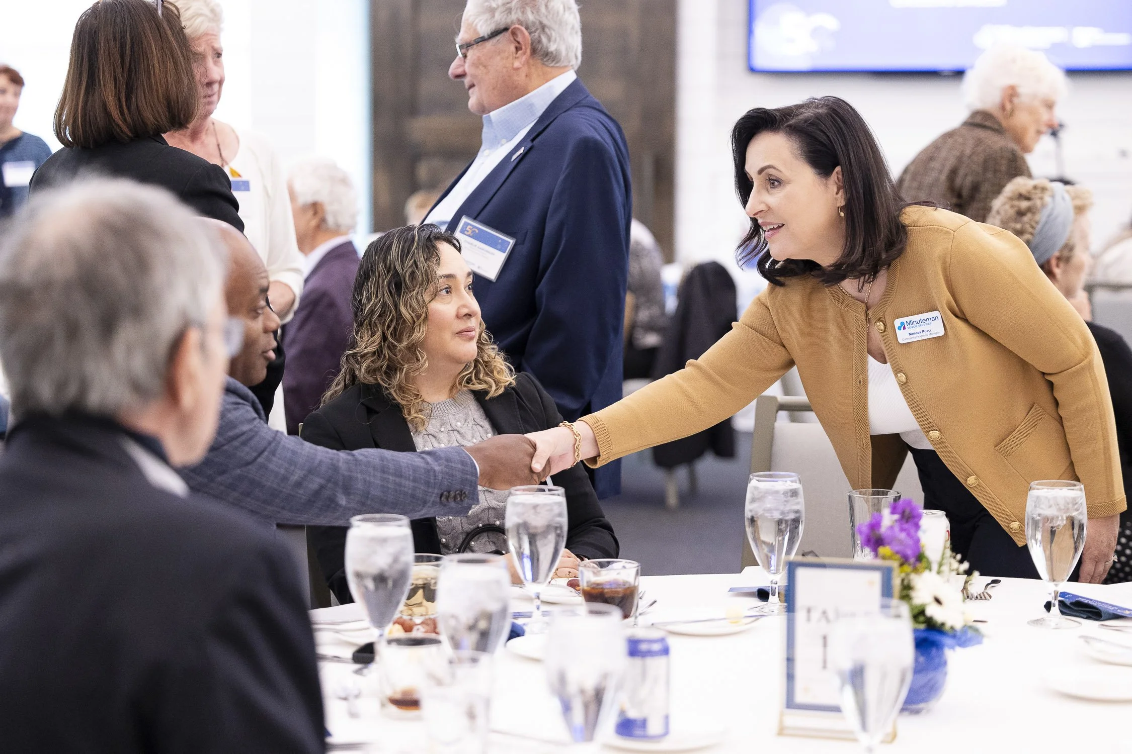 A woman shaking hands with a seated woman at a formal event in Boston.