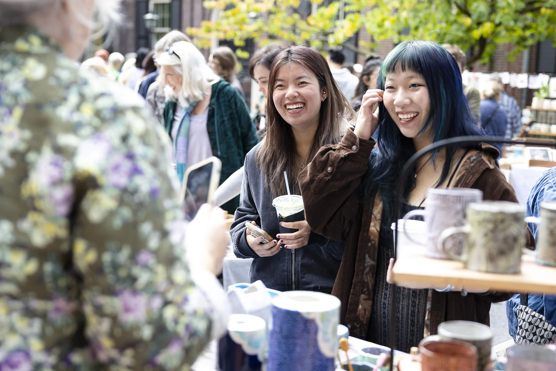 Two young women smiling at a market stall, one with turquoise hair and a brown jacket, the other with long brown hair and a dark hoodie, holding drinks and shopping at an outdoor market.