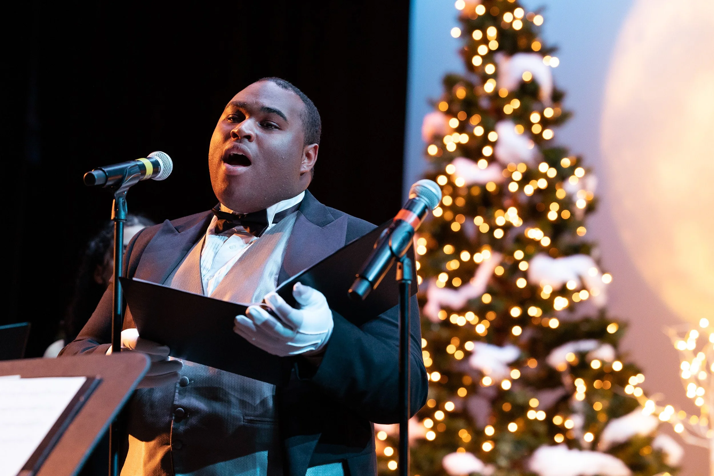 A male singer in a tuxedo singing into a microphone as part of a Christmas performance at Northeastern University.