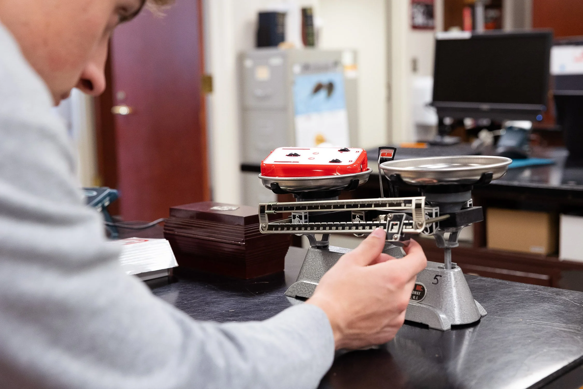 A person weighing a small red object on a balance scale in a college laboratory in Boston.