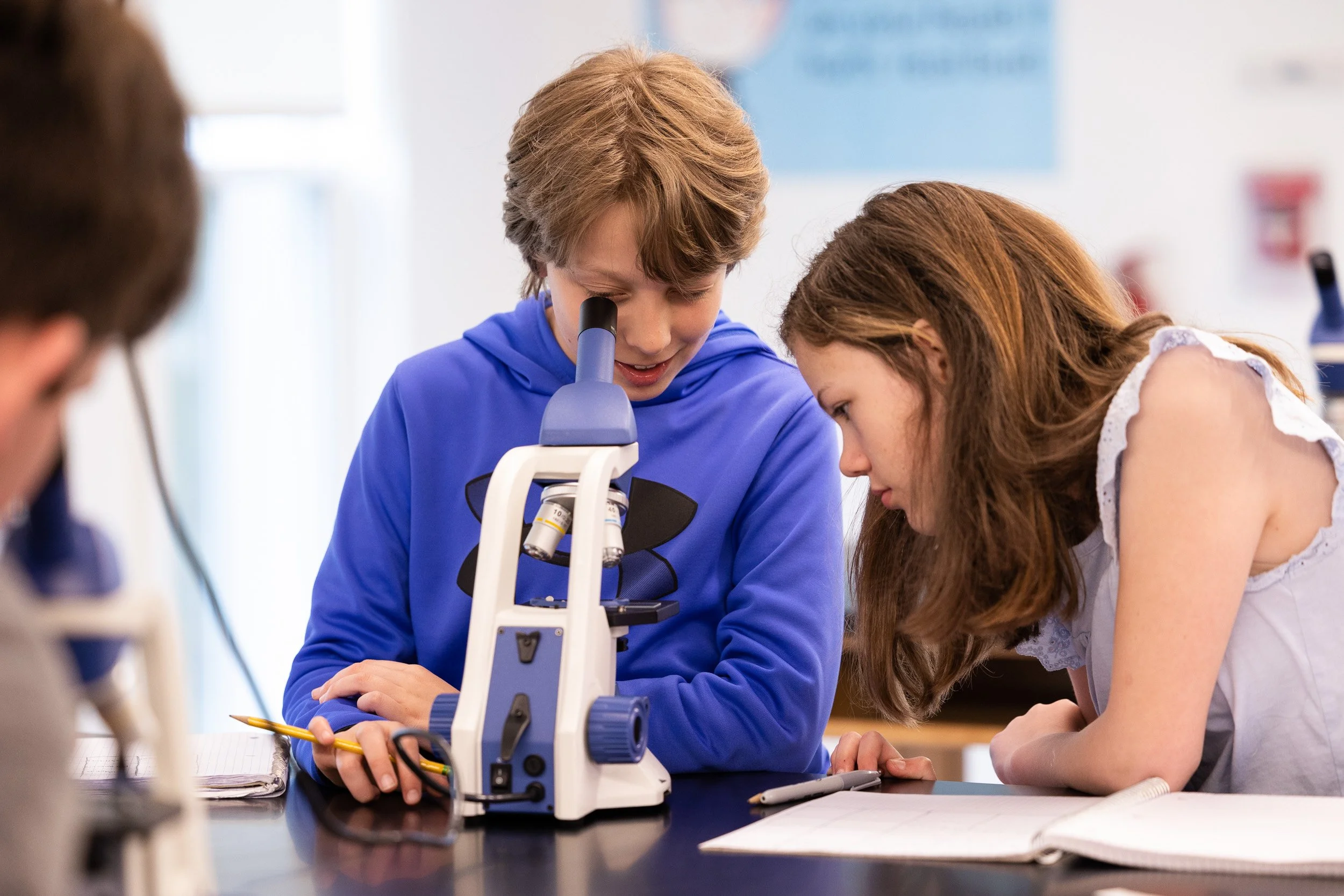Two middle school students, a boy and a girl, observe a microscope in a classroom at Charles River School.