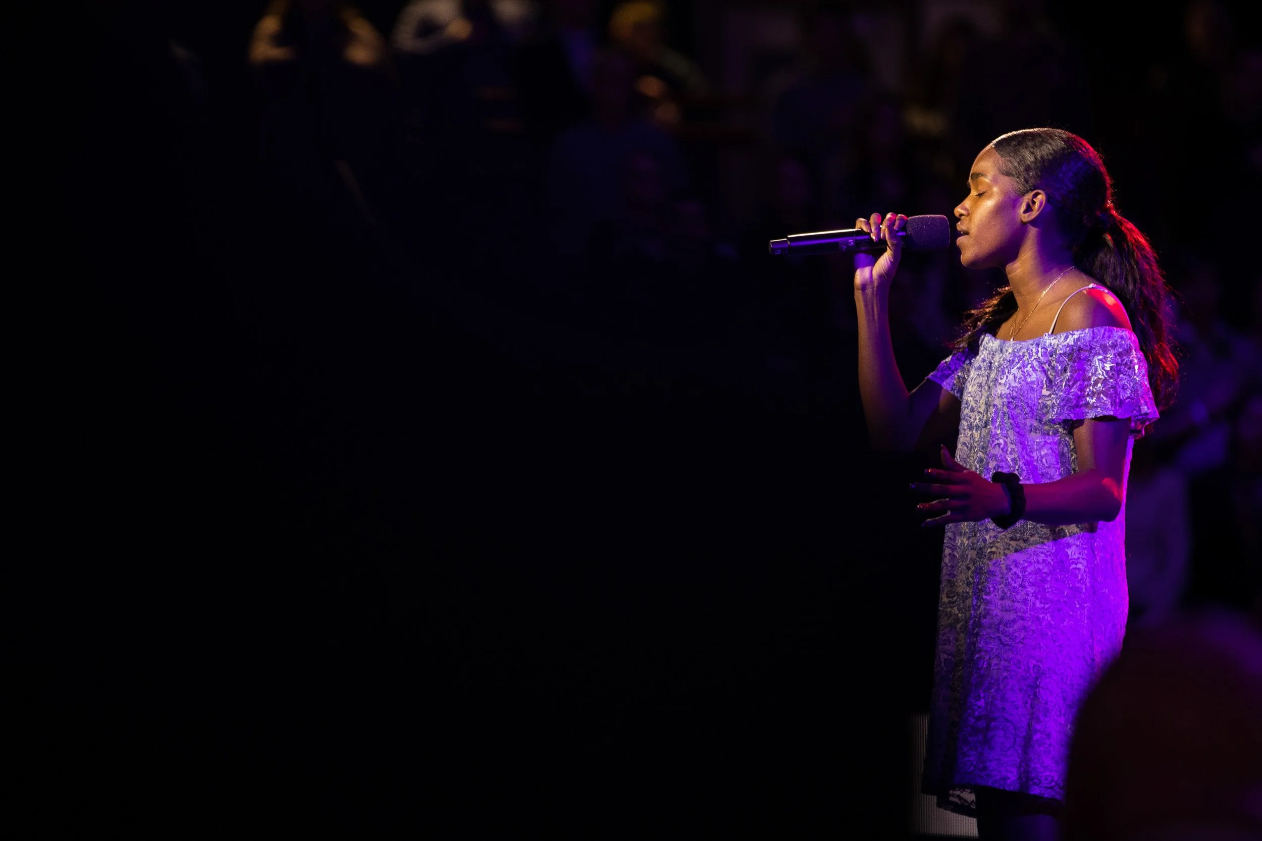 A young woman singing into a microphone on stage, with dark background and purple lighting.