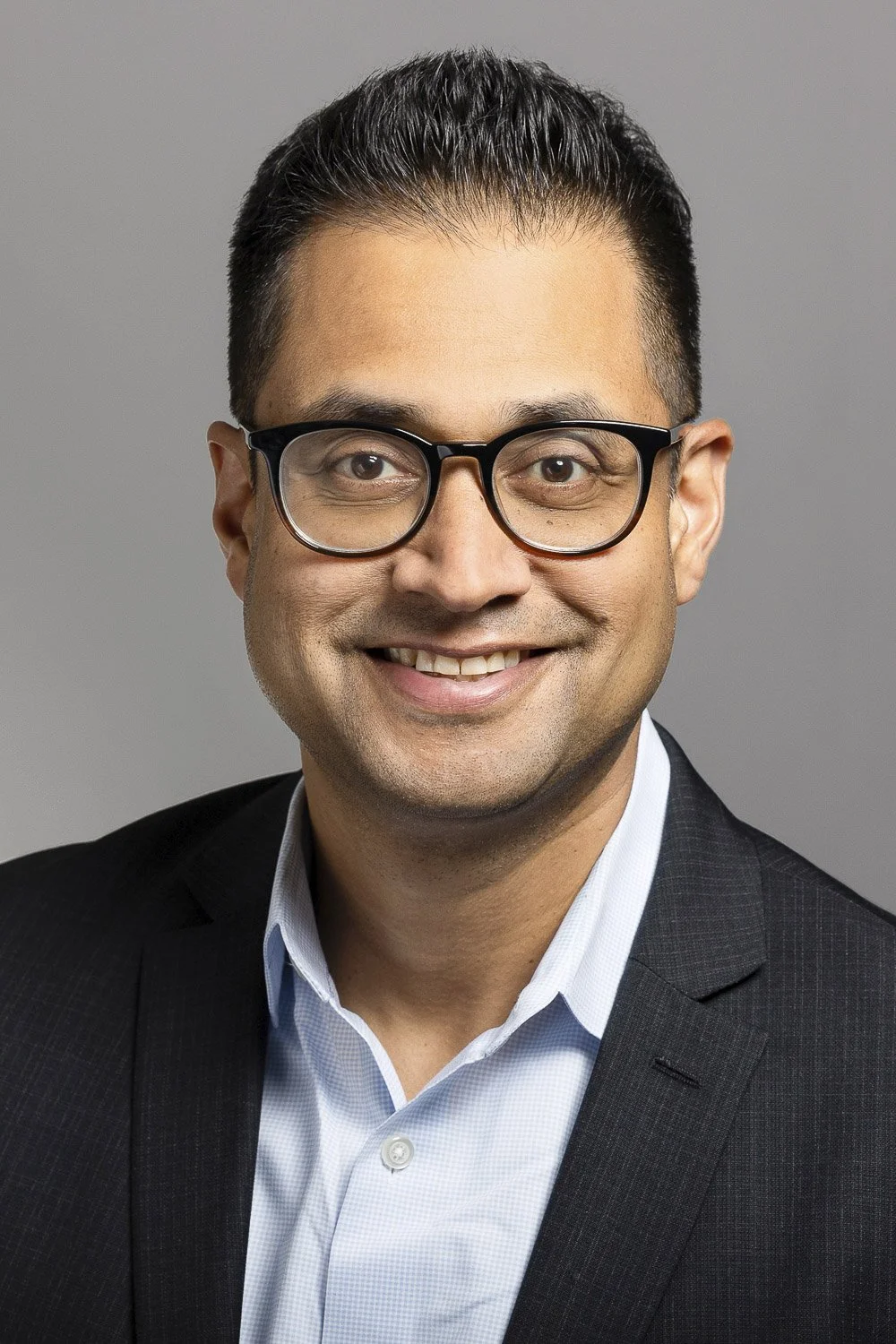 Corporate headshot of a smiling man wearing glasses, a light blue dress shirt, and a dark suit jacket against a gray background at a hotel in Boston.