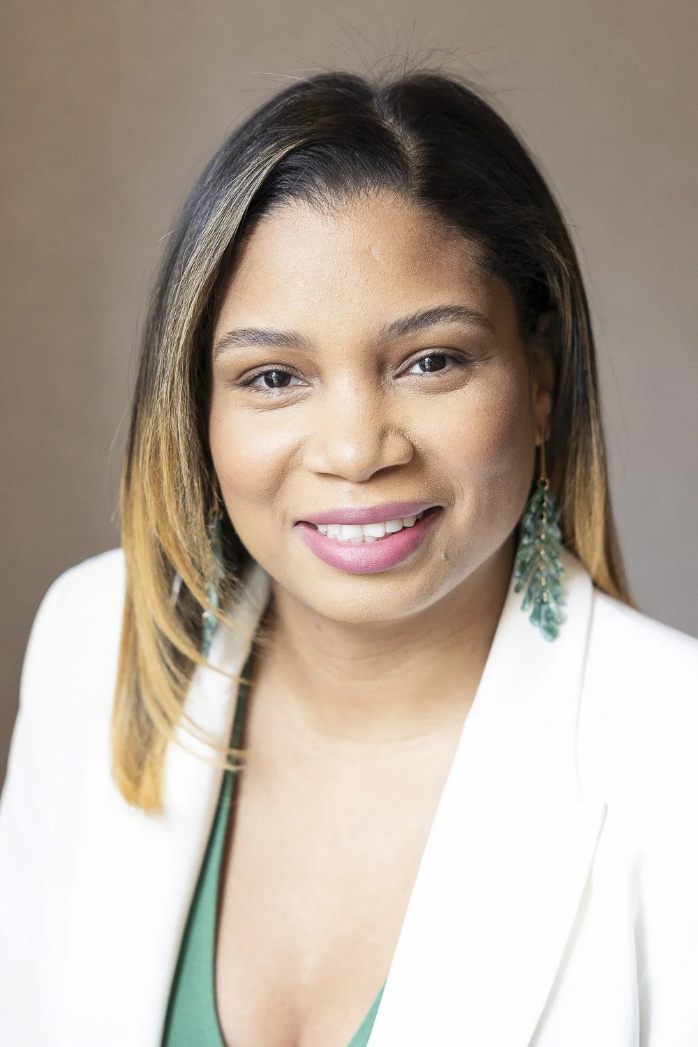 A woman with shoulder-length, light brown hair, wearing a white blazer and green top, smiling against a plain brown wall.