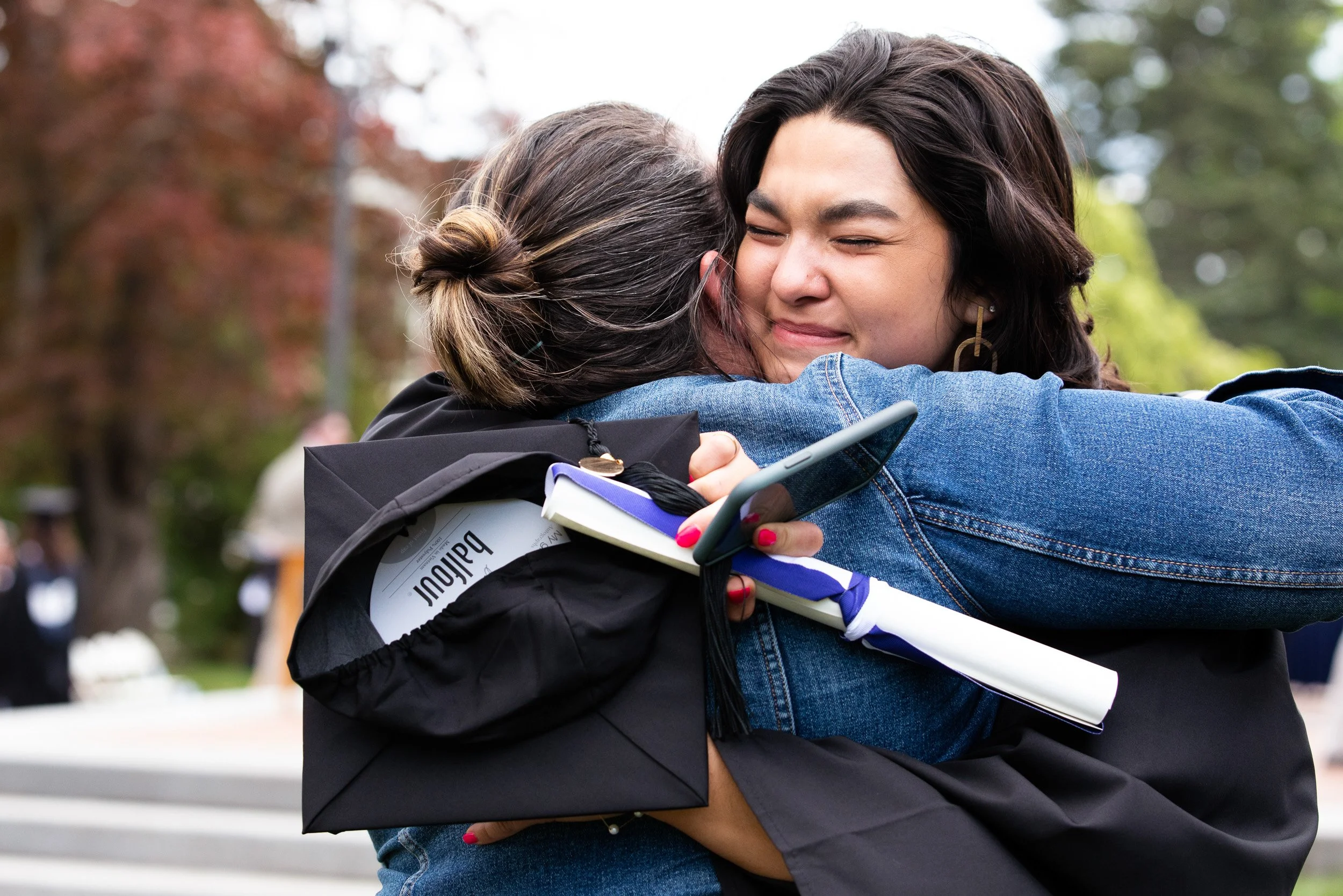 Two women hugging outdoors after a college graduation celebration, one in a graduation cap and gown.