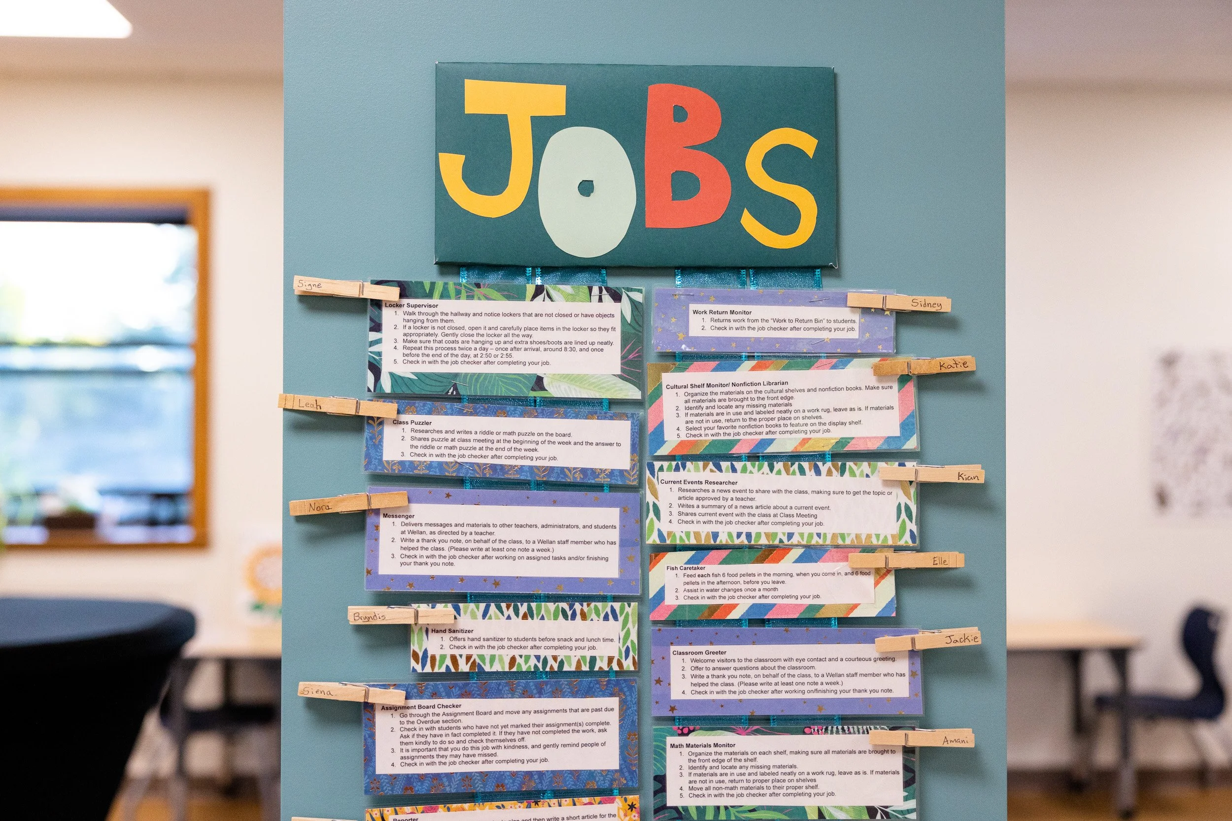 Montessori classroom bulletin board with a sign titled 'JOBS' and various job descriptions attached with clothespins, tape, and labels. 