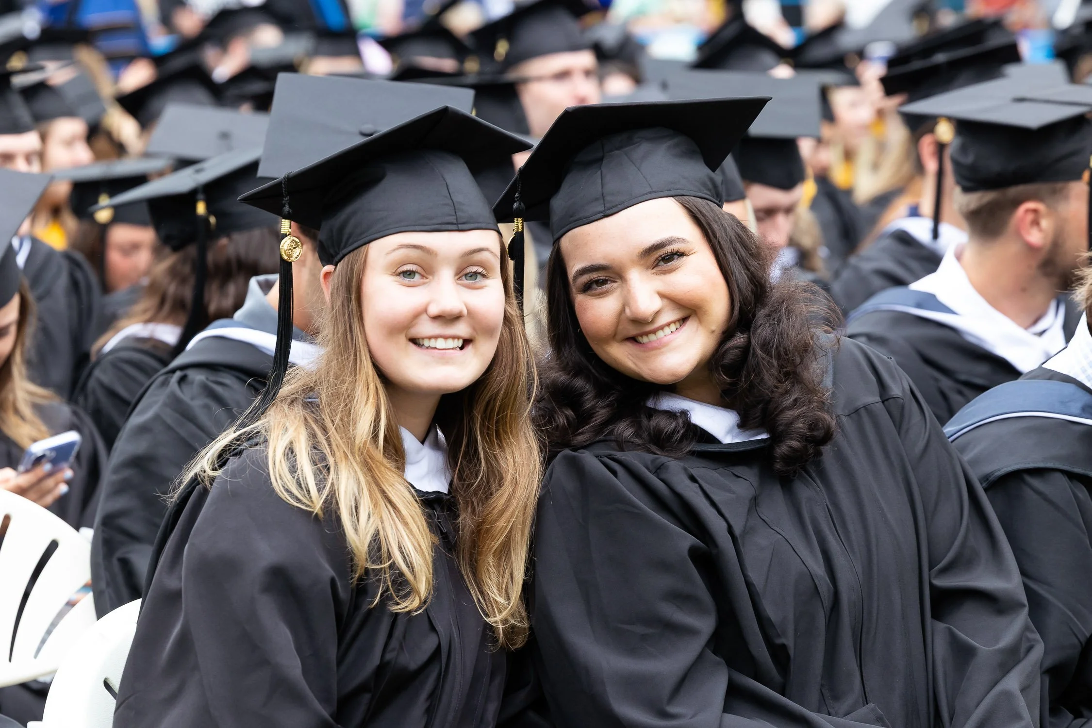 Two women in graduation caps and gowns smiling at an outdoor graduation ceremony.
