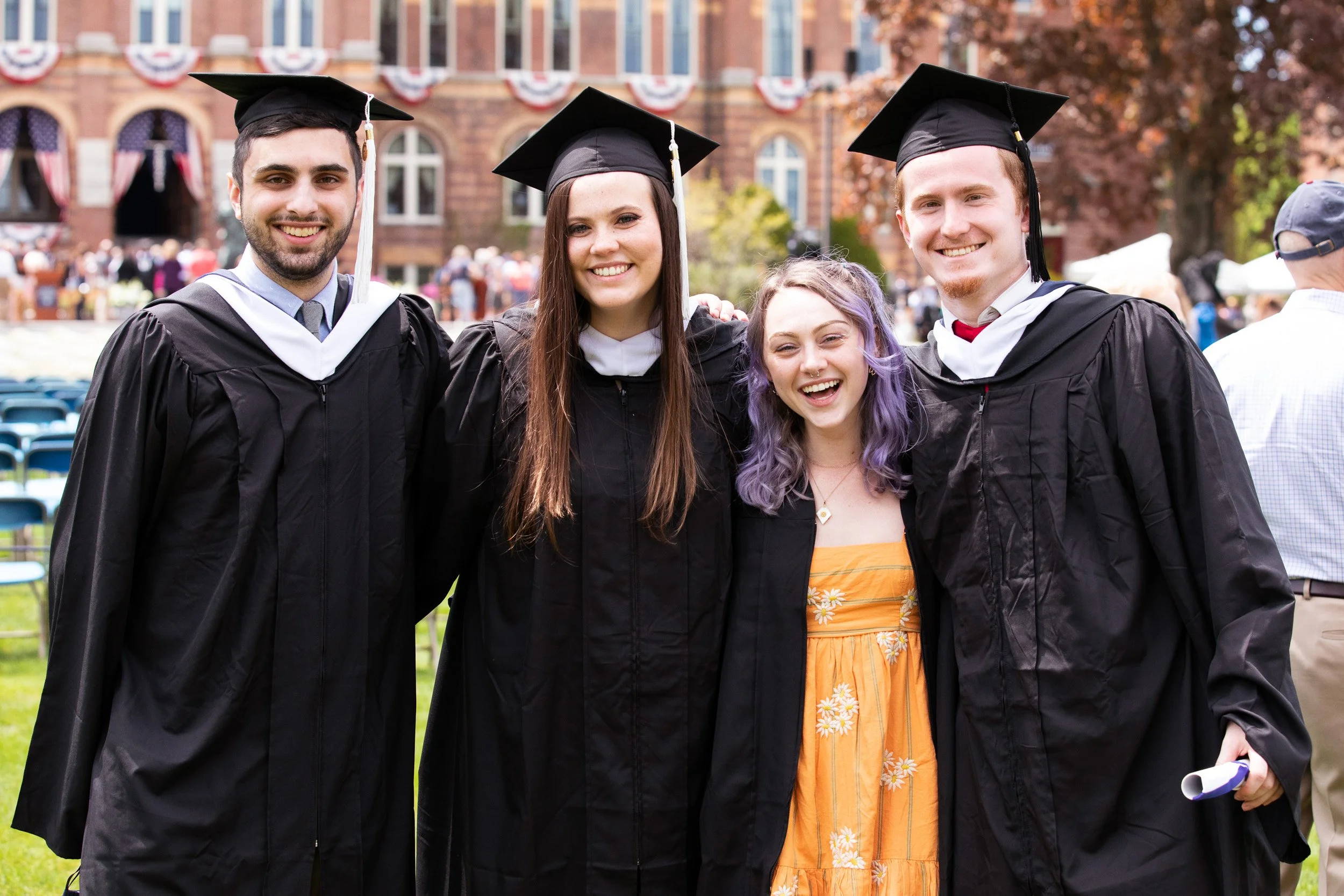 Four young adults in graduation caps and gowns smiling outdoors at a graduation ceremony at Saint Anselm College.