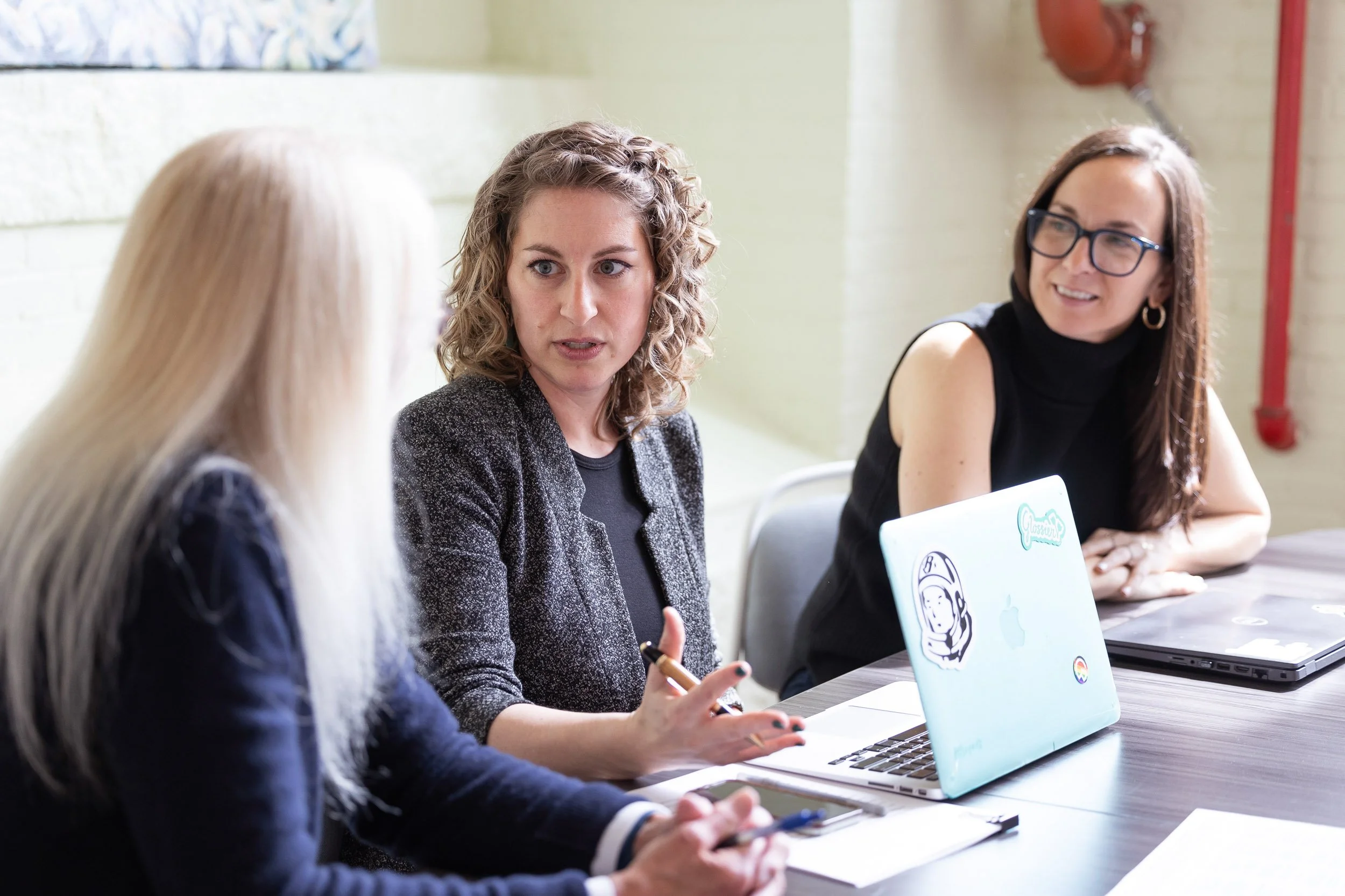 Three women with laptops and notebooks sitting at a table engaged in a discussion in a bright office space in Massachusetts.