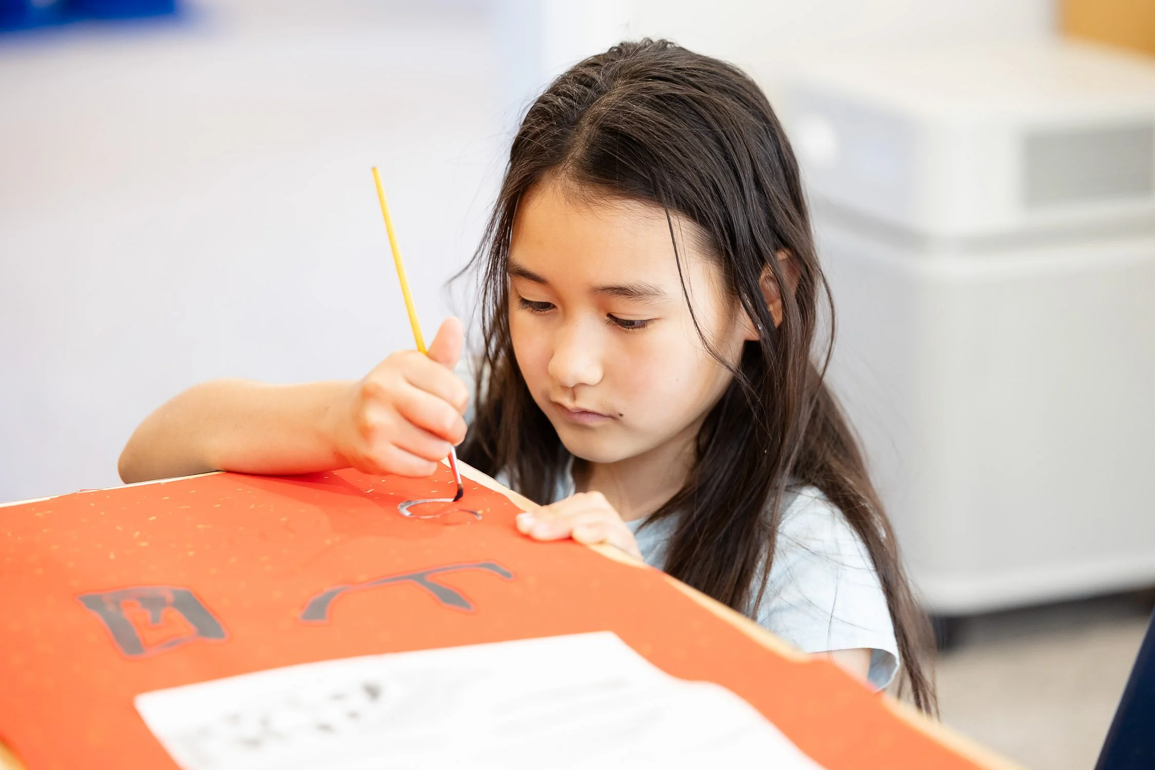 A girl creates letters on a red poster with black and gray paint, using a thin paintbrush.