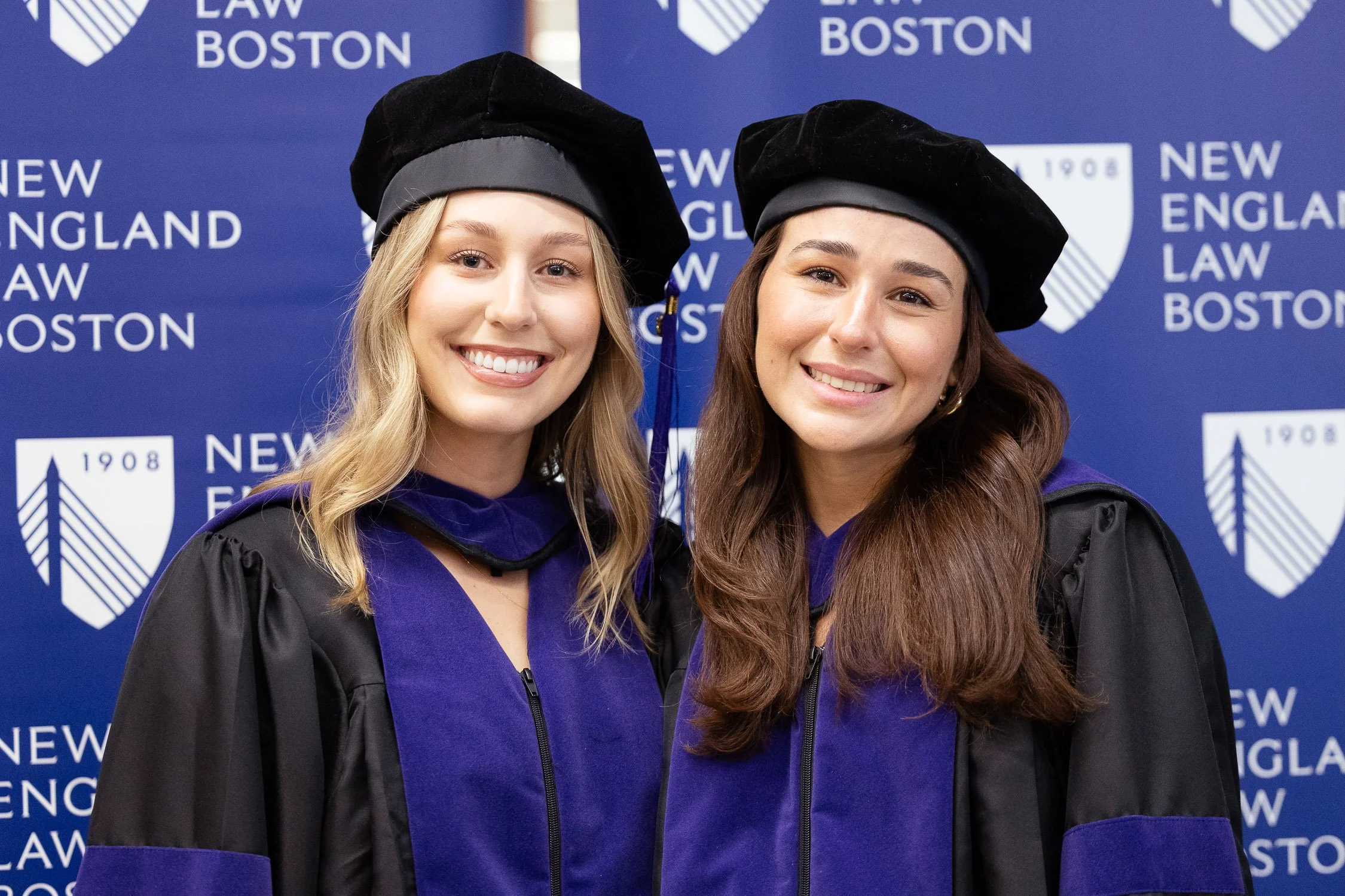 Two young women in graduation caps and gowns smiling at graduation ceremony with 'New England Law Boston' logo in the background.