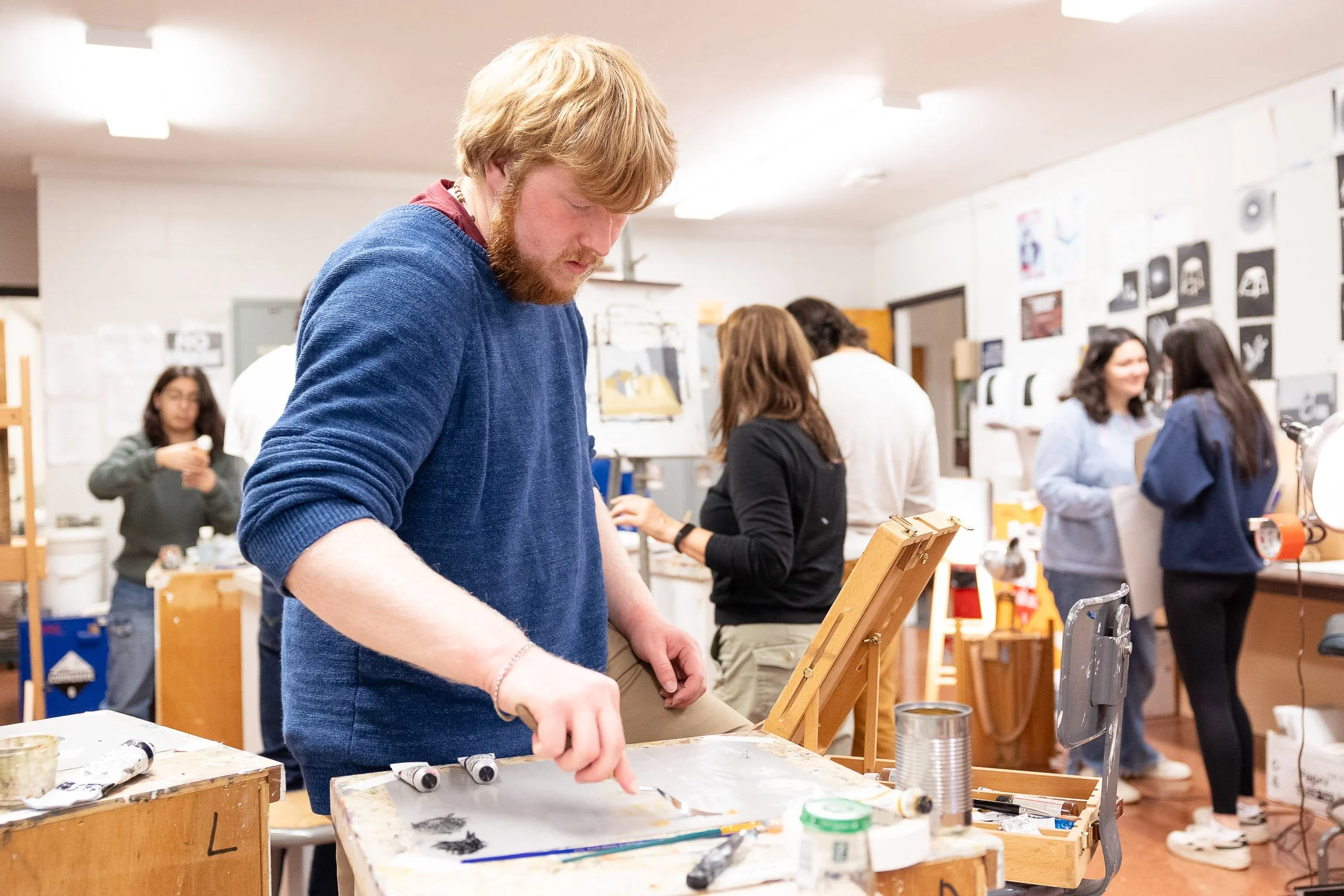A male college student working an art project in a classroom studio surrounded by other students. 