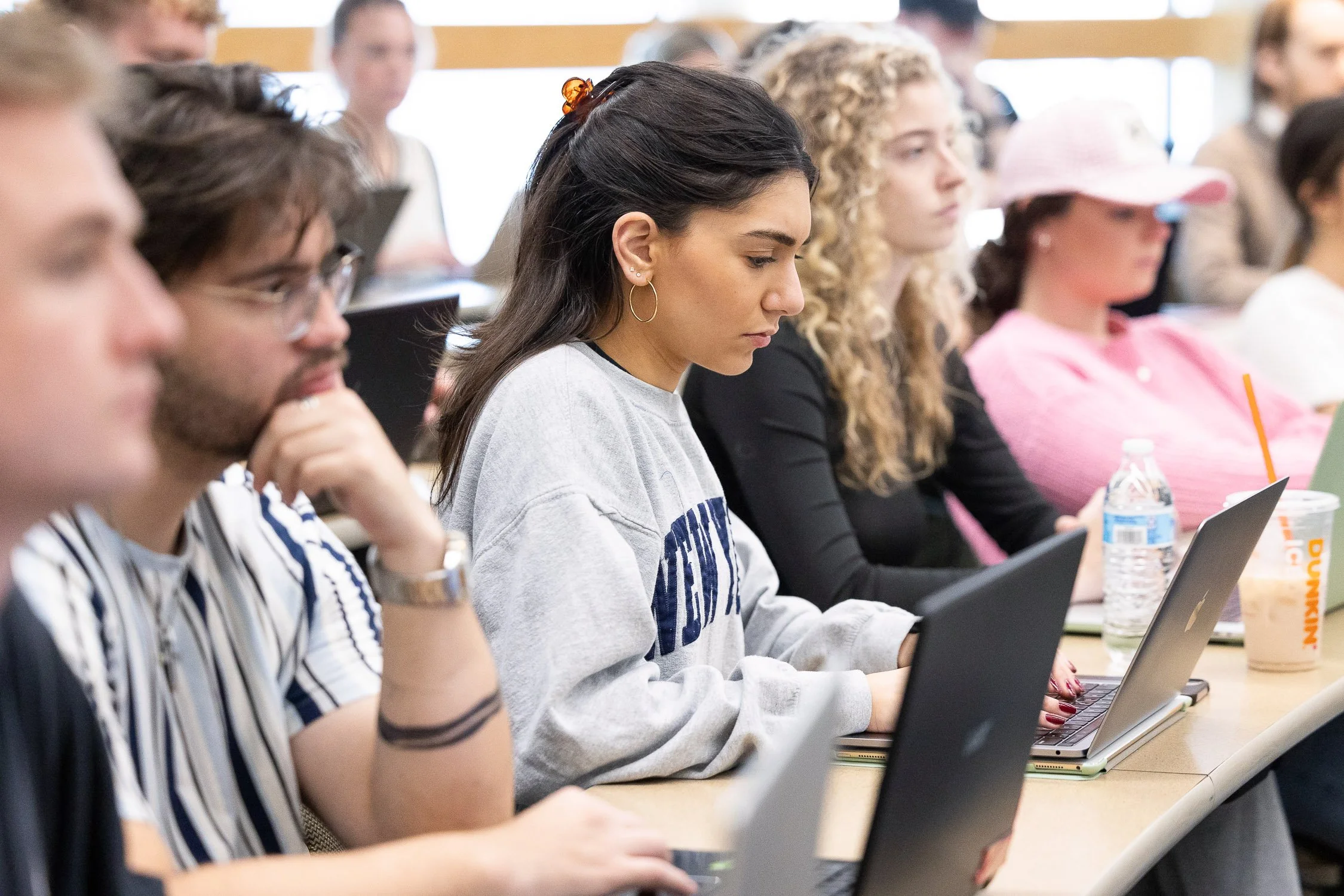 A group of students sitting at a classroom table, working on laptops, with focused expressions on a university campus in Boston.