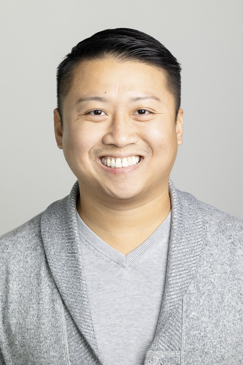 Portrait of a smiling man with short black hair wearing a grey sweater, standing against a plain light-colored background.
